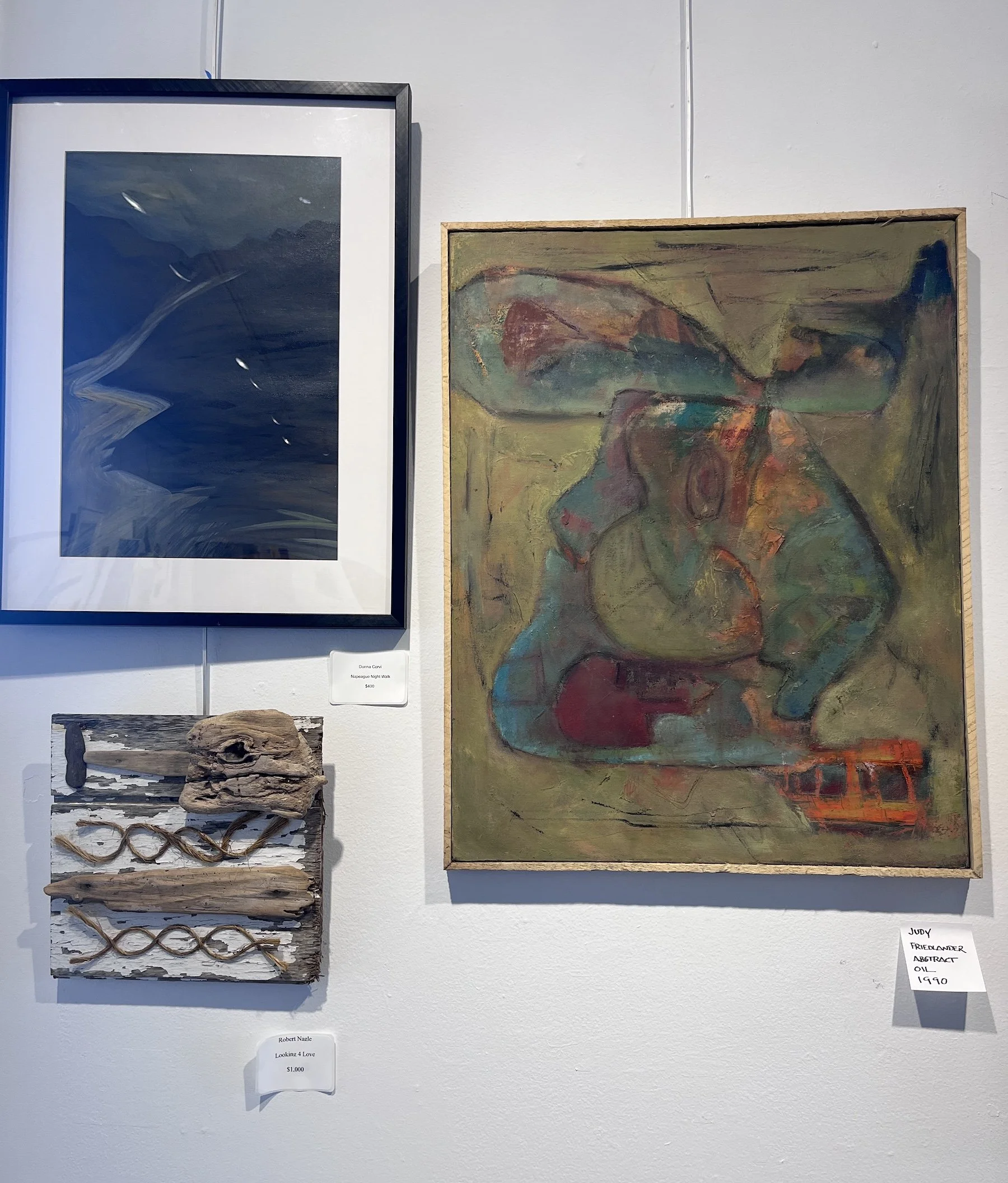 Art display on a white gallery wall featuring two paintings and a mixed media piece, with small labels indicating artist and title. The left artwork is an abstract painting with dark blues and swirling white streaks framed in black and white. The right artwork is an abstract painting with earthy greens, reds, and blues in a wooden frame. The mixed media piece below consists of driftwood and rope arranged on a painted wooden board.