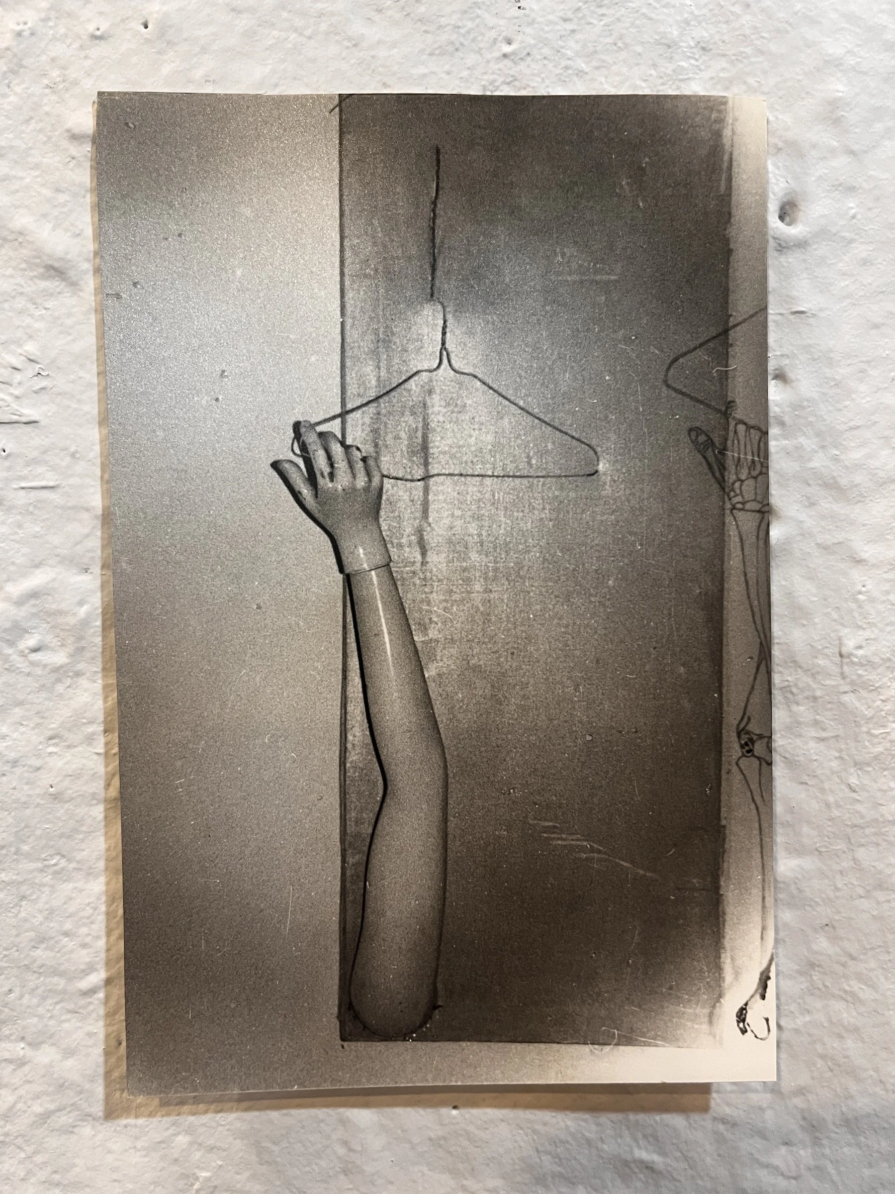 Black and white photograph of a hand holding a wire hanger with a coat hanger shape, on a textured wall background.