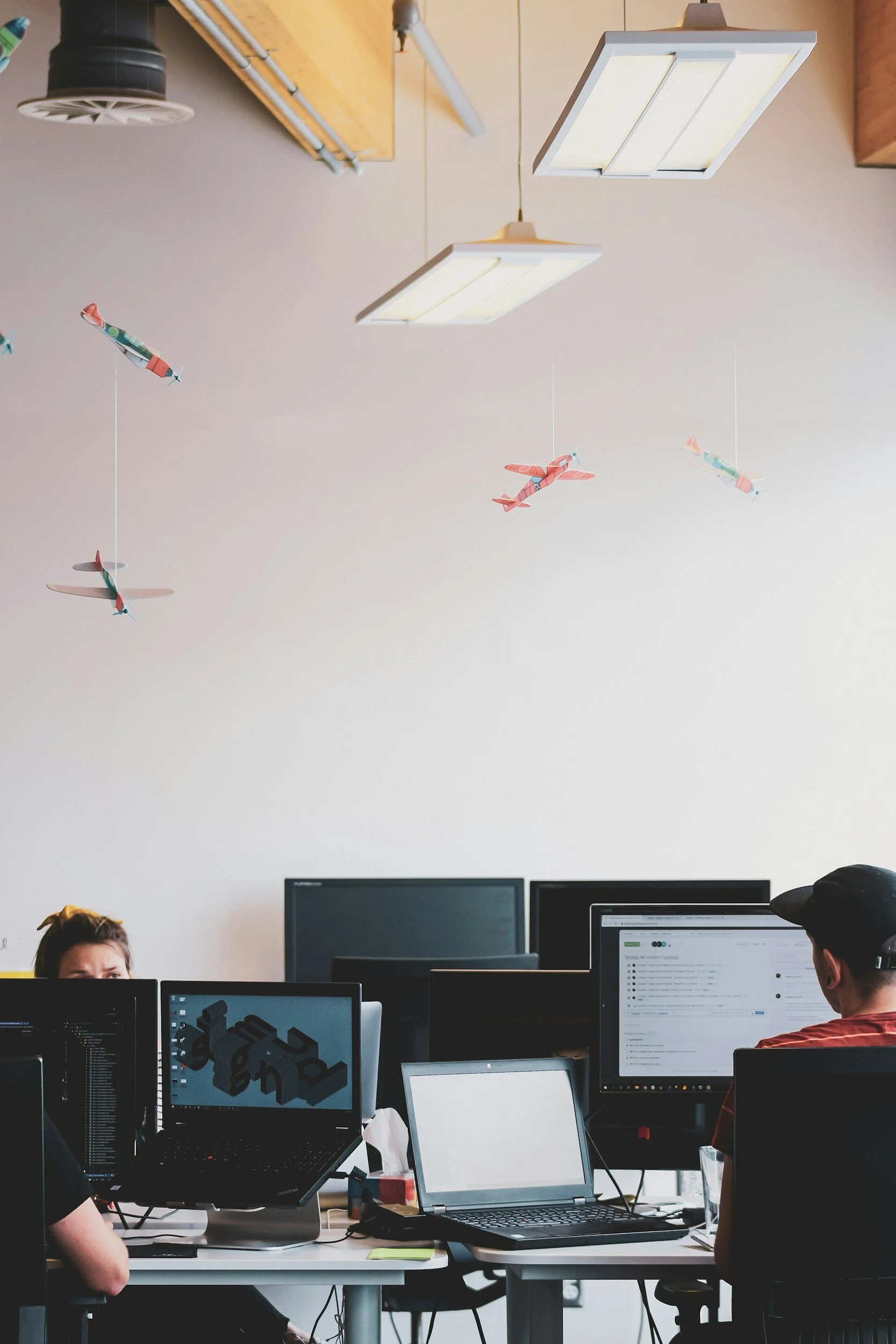 An office workspace with multiple computer monitors, some displaying coding or 3D modeling software. In the background, there are people working at desks, and the ceiling has hanging airplane-shaped decorations along with ceiling lights and an air vent.