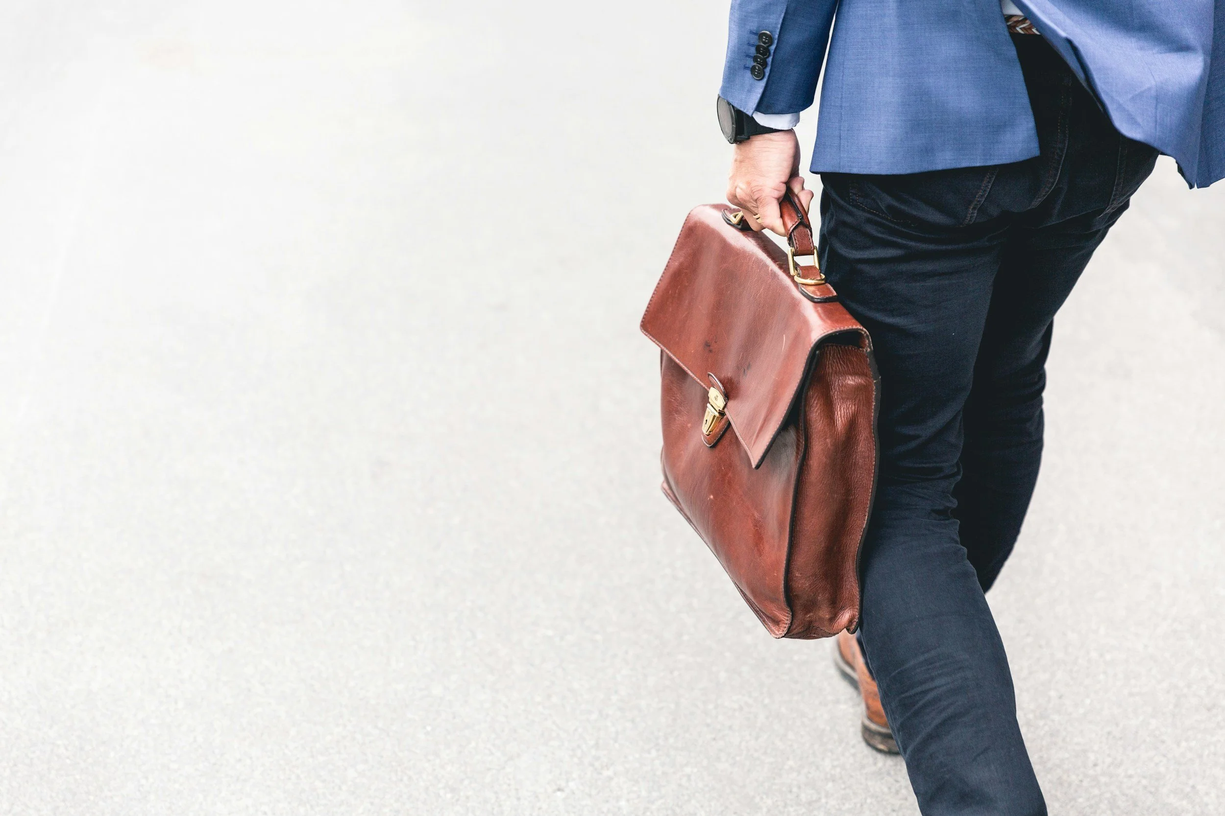 A man in a blue suit jacket, dark jeans, and brown shoes is walking on a light-colored pavement, carrying a brown leather briefcase.