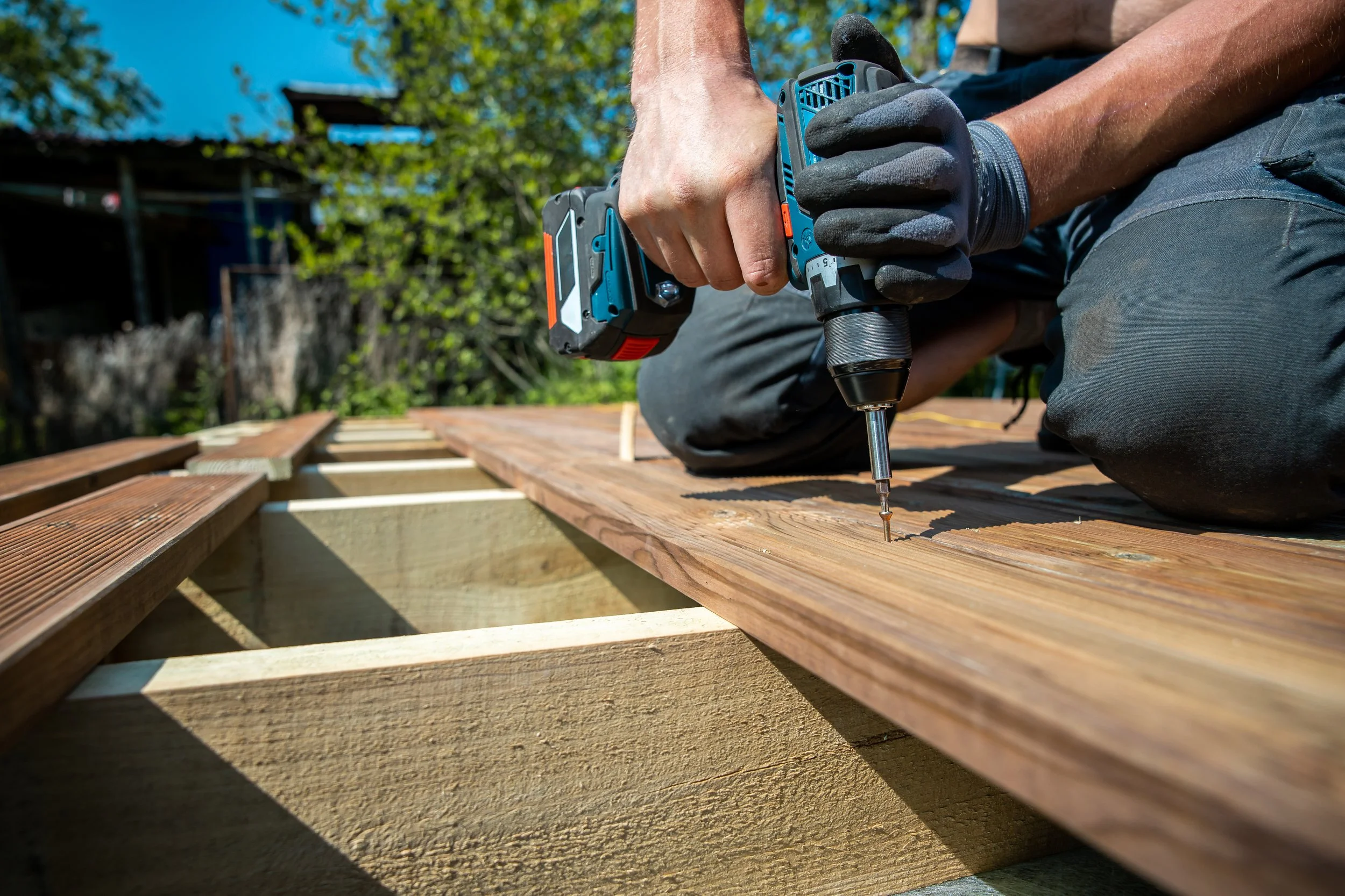 A person working on a wooden deck using a cordless drill, wearing gloves, with a background of trees and a house under a blue sky.