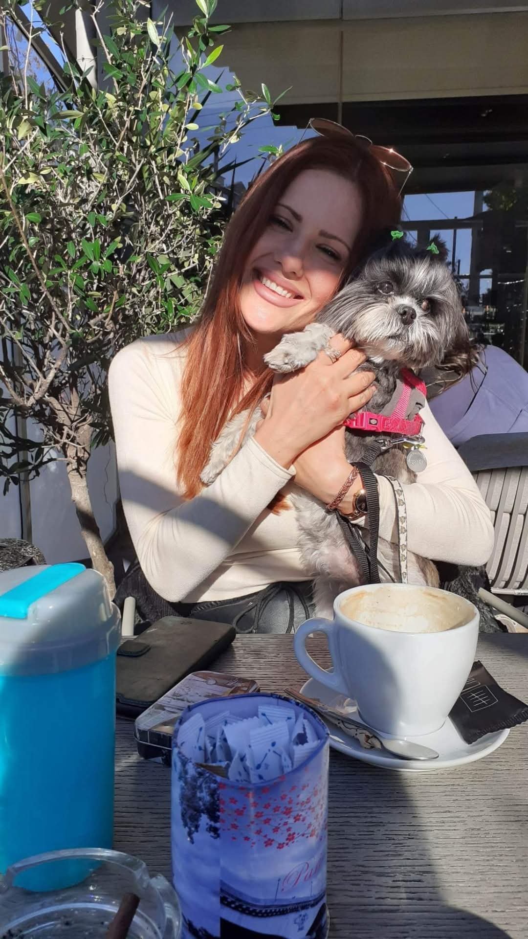 A woman with long red hair smiling and holding a small gray and white dog with a pink harness, sitting outdoors at a table.