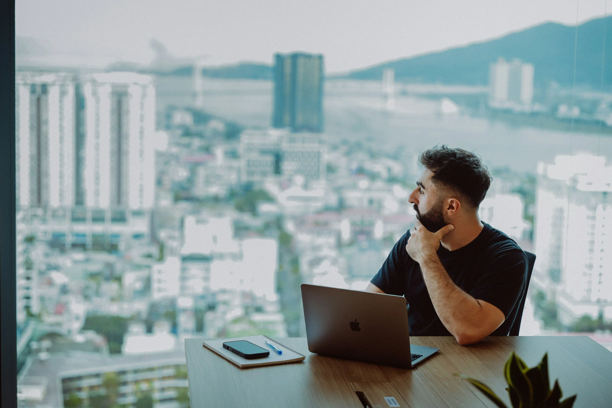Man sitting at a desk with a MacBook, smartphone, notebook, and pen, looking thoughtfully out a large window at a cityscape with high-rise buildings and distant mountains.
