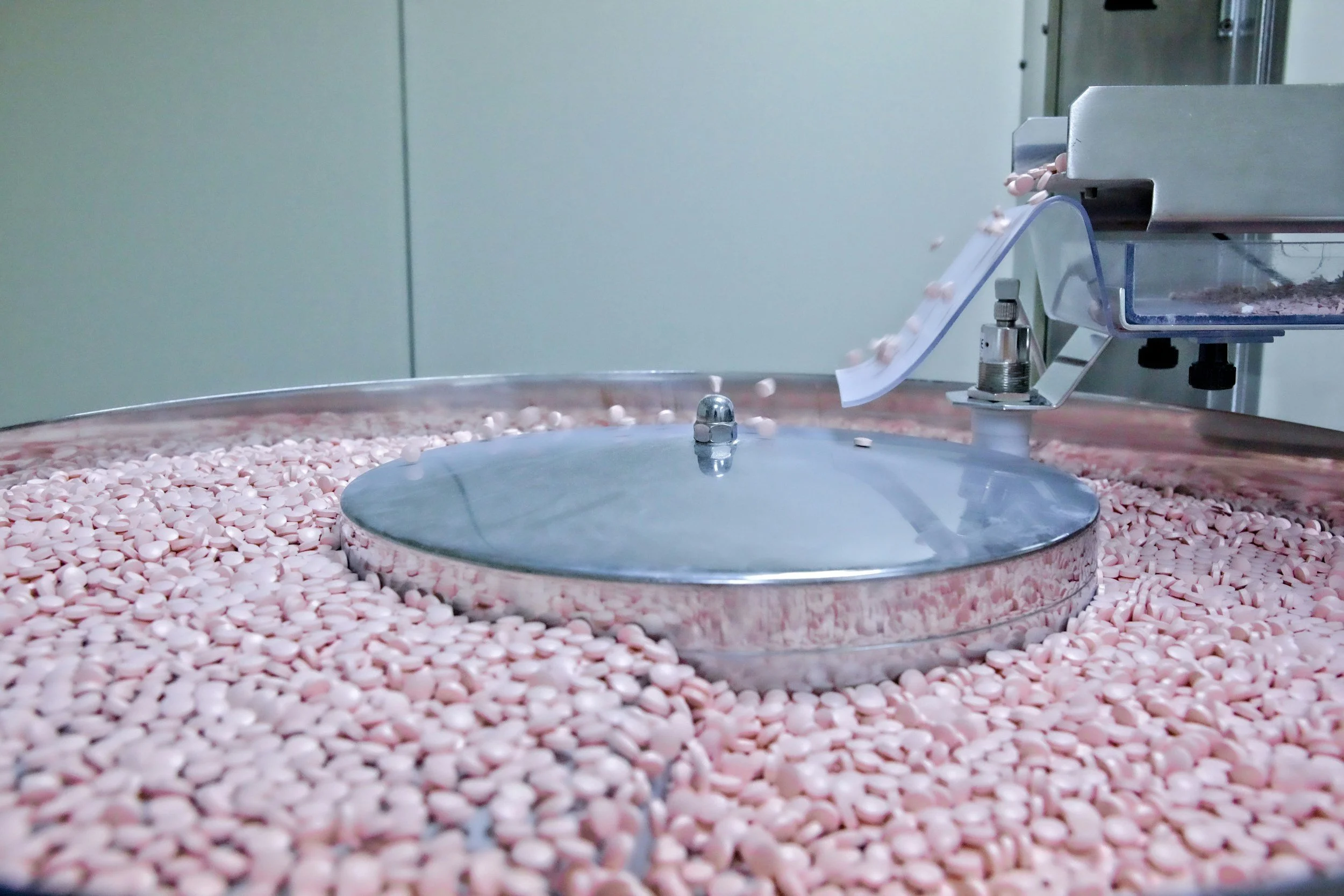 A pharmaceutical pill manufacturing machine dispensing small pink pills onto a conveyor belt.