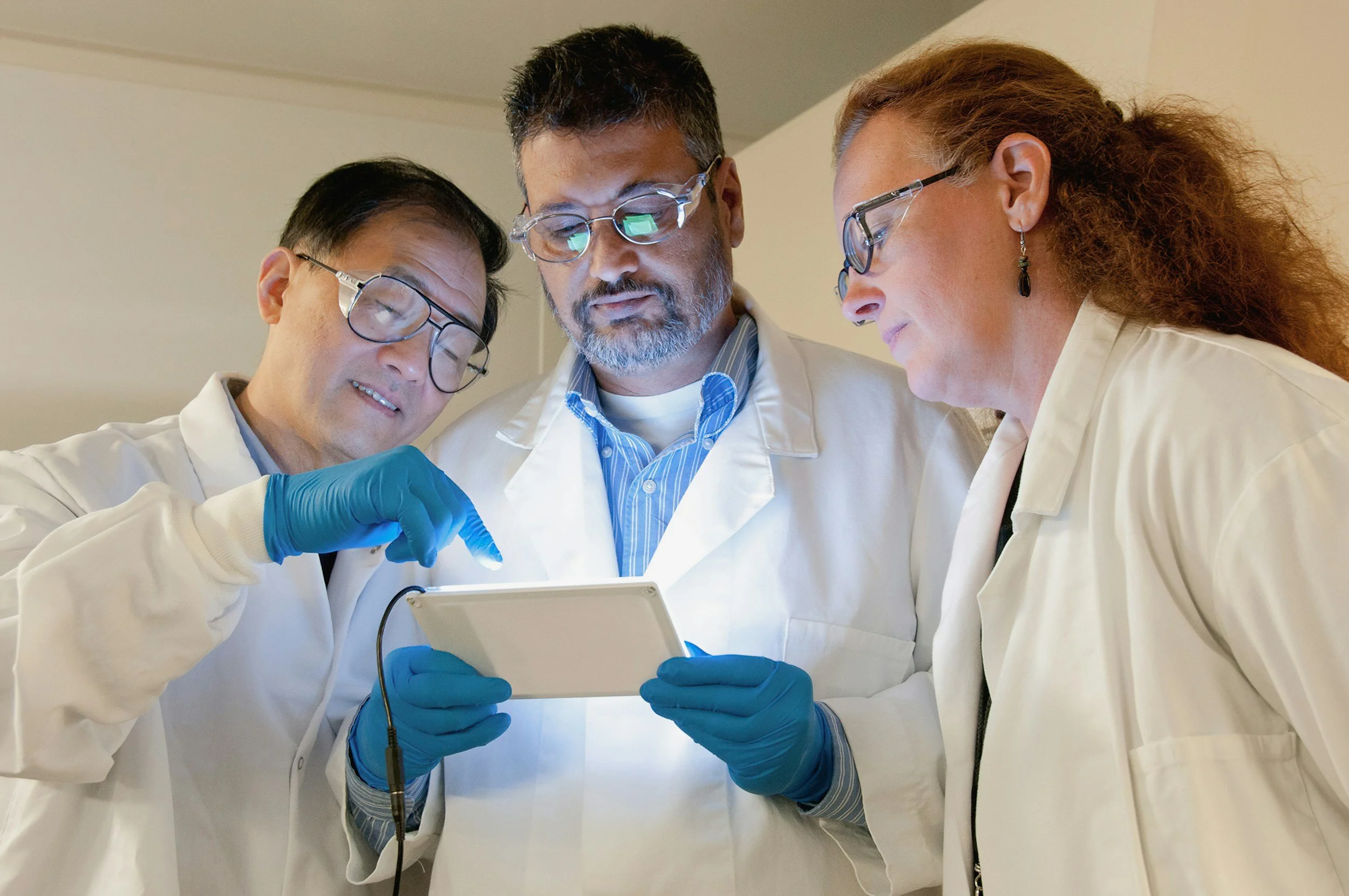 Three doctors in white coats and gloves gathered around looking at a tablet device.