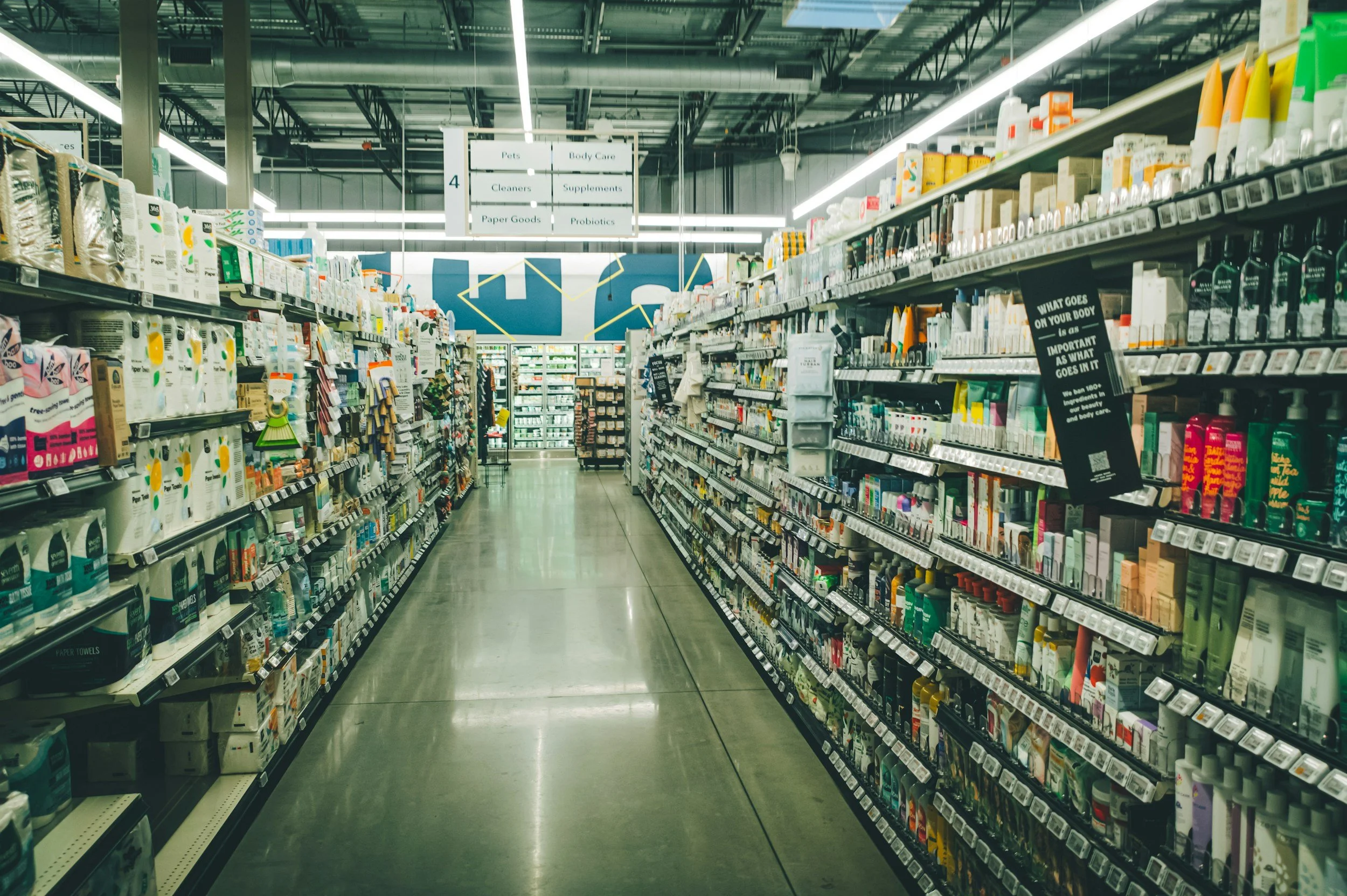Aisle in a pharmacy or grocery store with shelves stocked with personal care, health, and cleaning products. Overhead signs guide shoppers to pet supplies, body care, cleaners, supplements, paper goods, and probiotics. The aisle has bright, even lighting and a polished concrete floor.