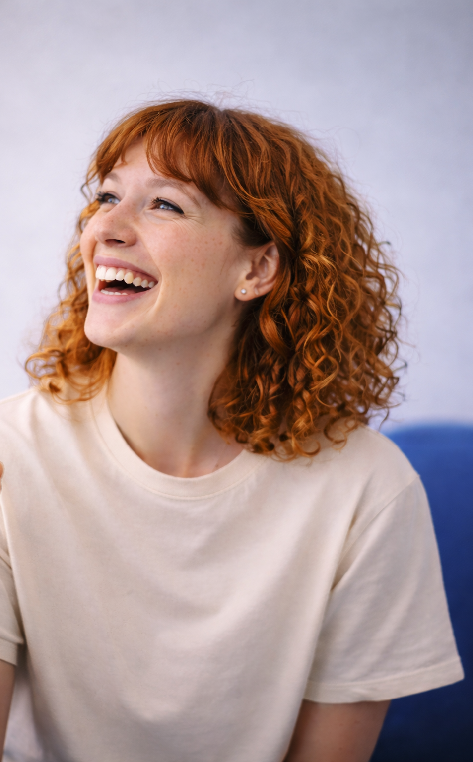 A young woman with curly red hair, light skin with freckles, laughing with her mouth open, wearing a beige t-shirt, sitting against a neutral background.
