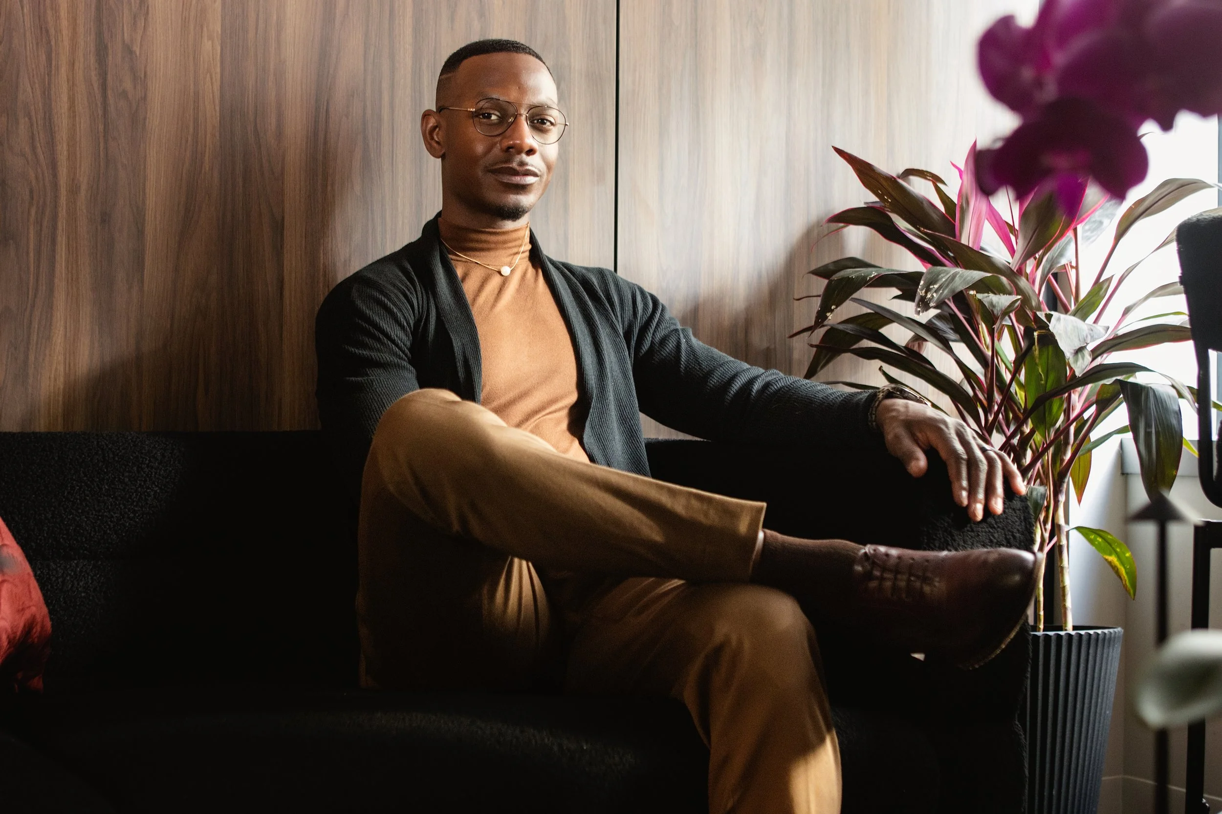 A man sitting on a black sofa, wearing glasses, a brown turtleneck, a black cardigan, and tan pants, with a wooden wall and a large plant nearby