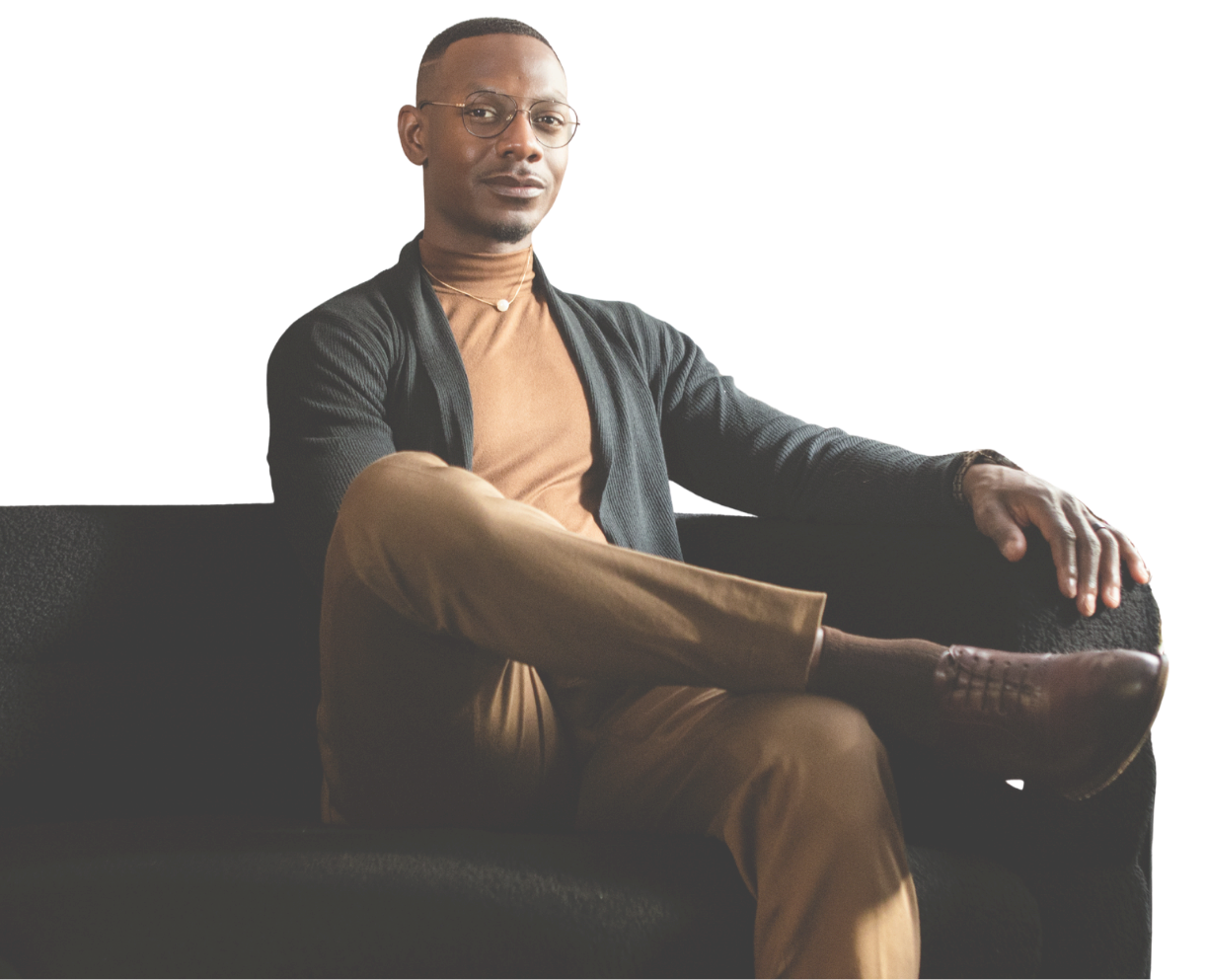A man sitting on a black sofa, wearing glasses, a brown turtleneck, brown pants, a black cardigan, and brown shoes, with a neutral expression.