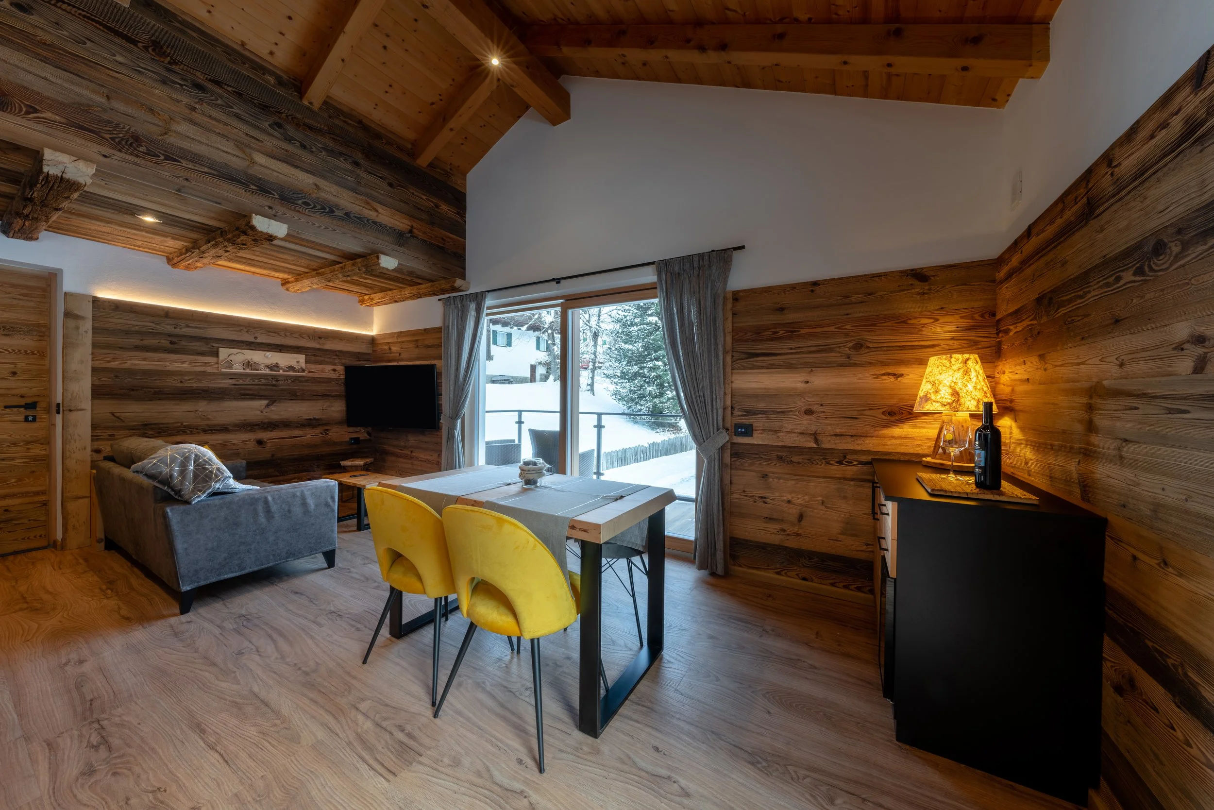 Interior di un soggiorno in stile chalet con pareti e soffitto in legno, tavolo di legno con sedie gialle, divano grigio, televisore a parete e vista sulla neve attraverso una grande finestra con tende grigie, lampada accesa su un mobile nero.
