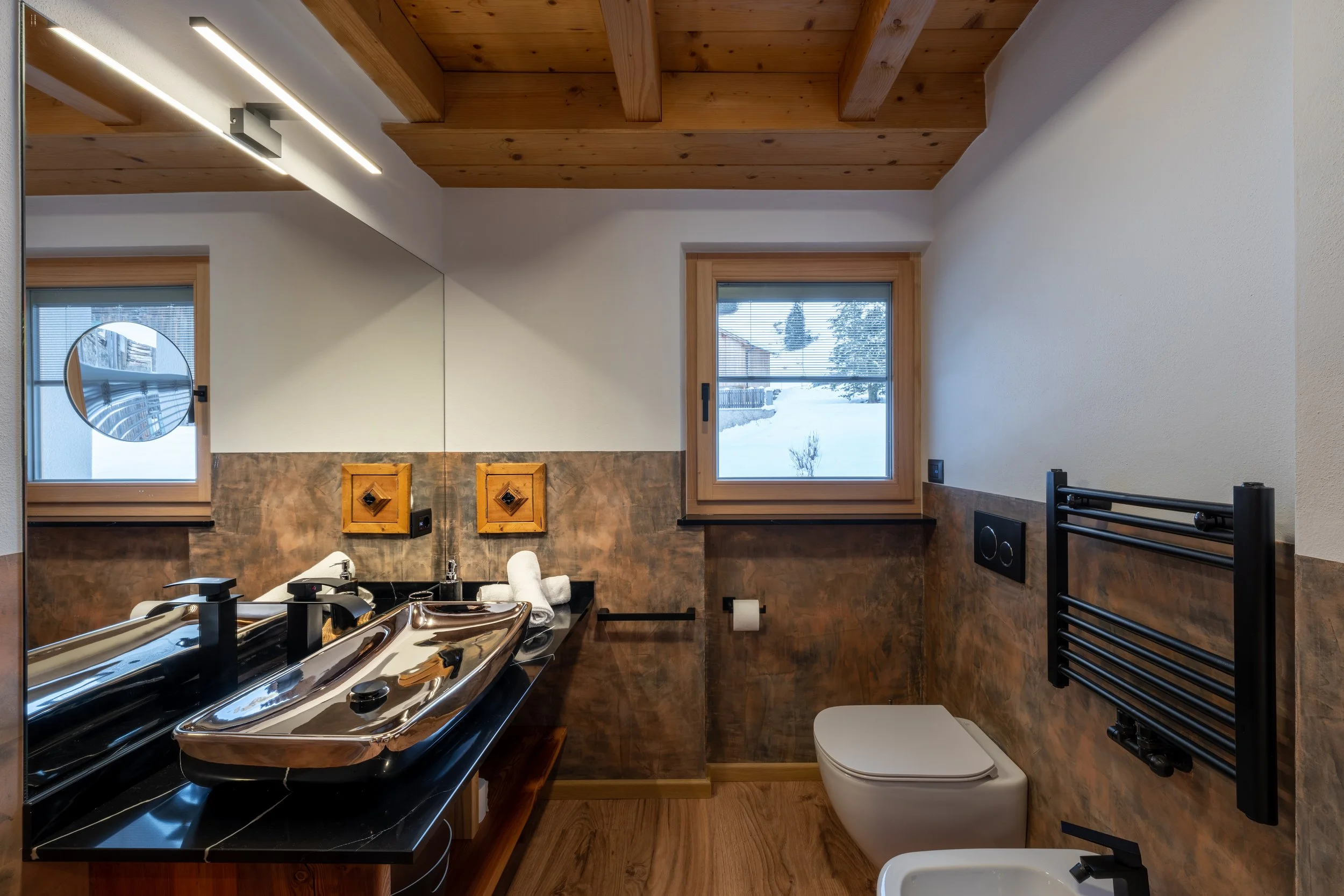 Bagno con lavabo in acciaio, specchio grande, finestra con vista sulla neve, pareti in pietra e soffitto in legno