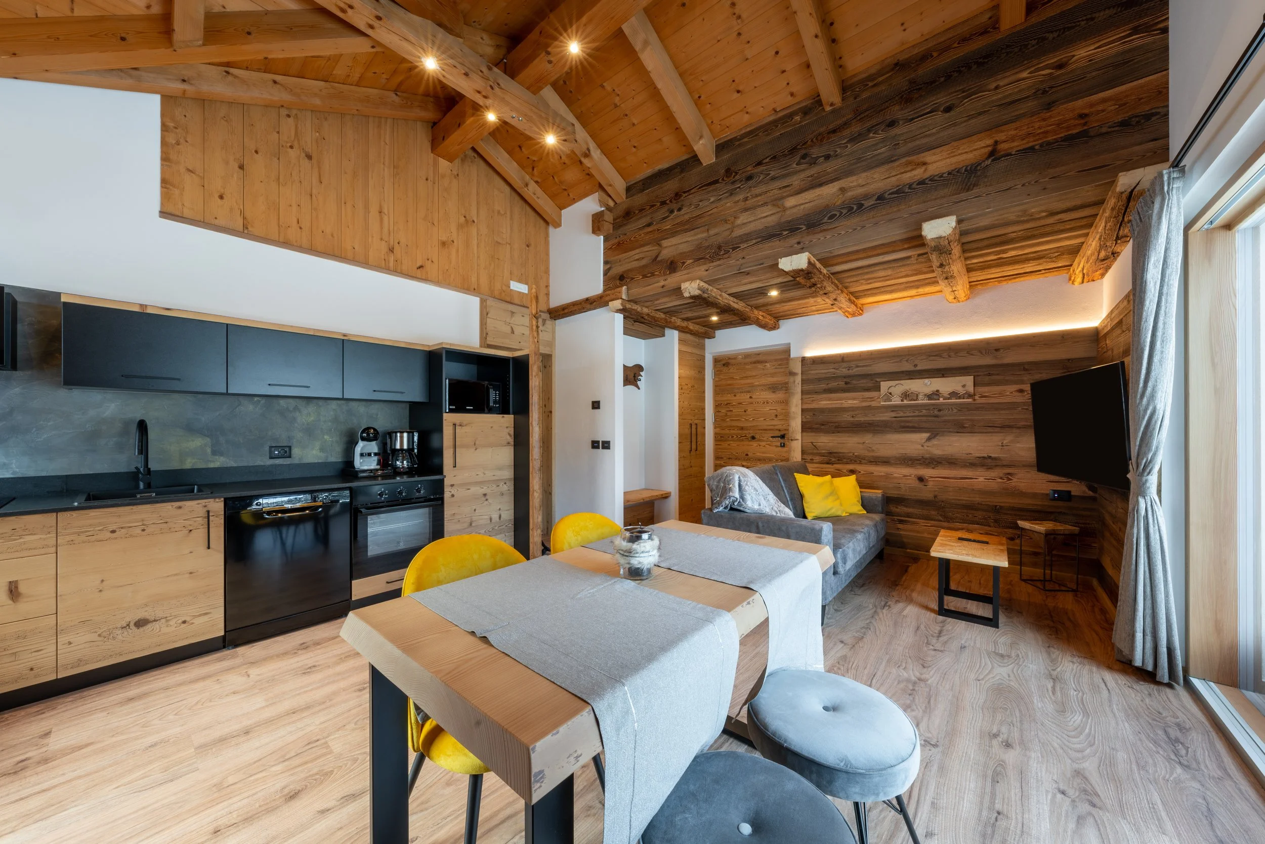 Interno di una casa in stile rustico con soffitto in legno, cucina moderna e sala con divano, tavolo e TV