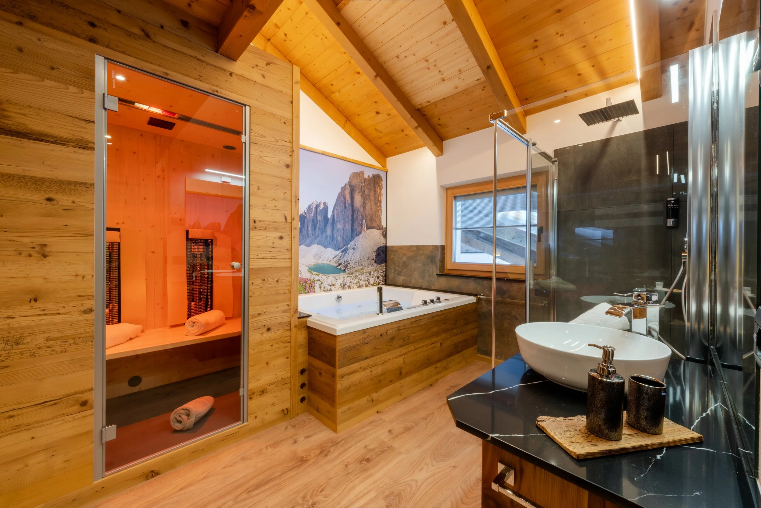 Bagno moderno con sauna in legno, vasca da bagno con vista montagna, doccia con soffione a pioggia, lavabo rotondo con accessories neri su superficie in marmo nero.