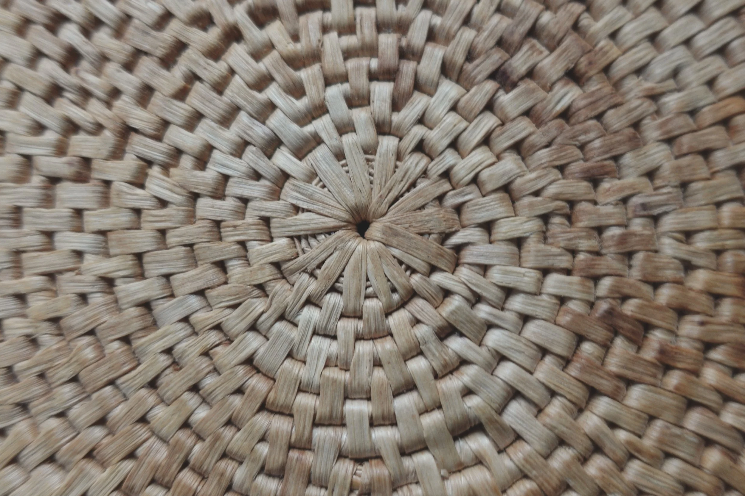 Close-up of a woven basket or mat made of natural fibers, showing an intricate circular pattern with a small hole at the center.