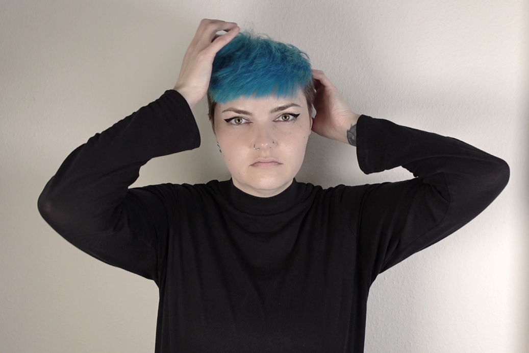 Person with short, blue and teal hair, wearing a black long-sleeve shirt, standing against a plain wall, holding her head with both hands, looking directly at the camera with a serious expression.