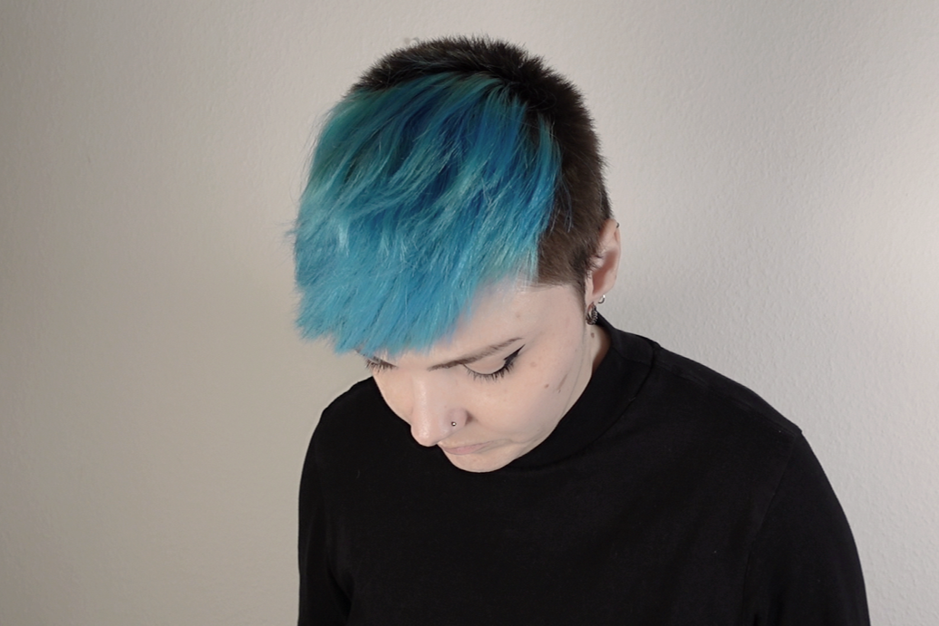Person with short black hair and a bright blue streak, wearing a black shirt, looking downward against a plain light-colored wall.