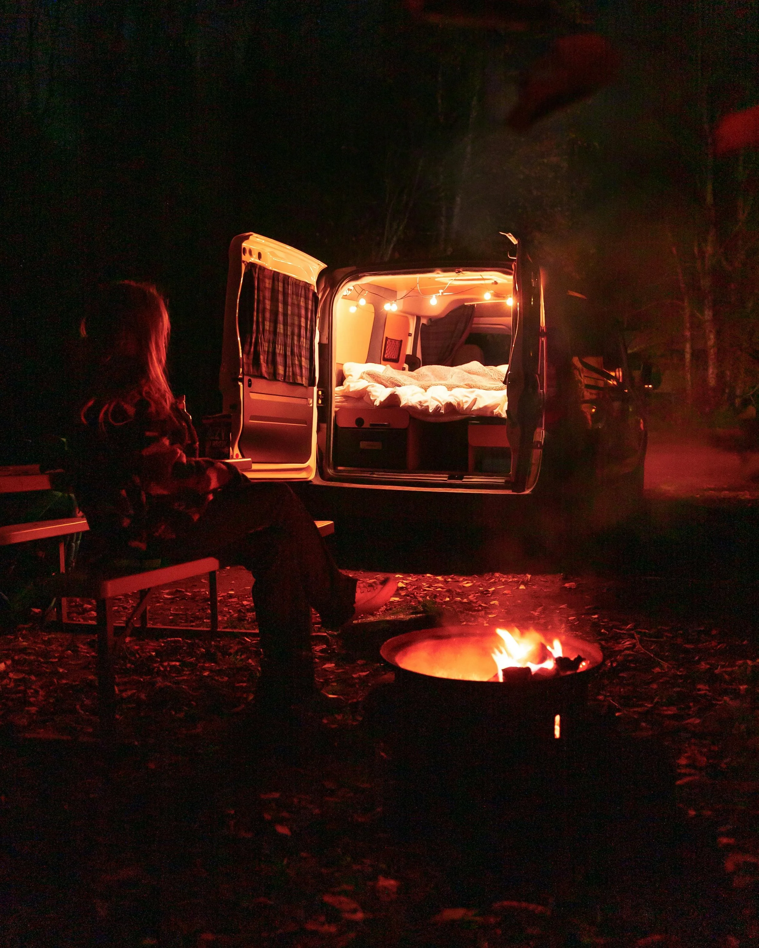 Camper met open deuren en sfeervolle verlichting, in de avond bij een kampvuur, in een rustige bosomgeving tijdens een solo retreat van een 45+ vrouw.