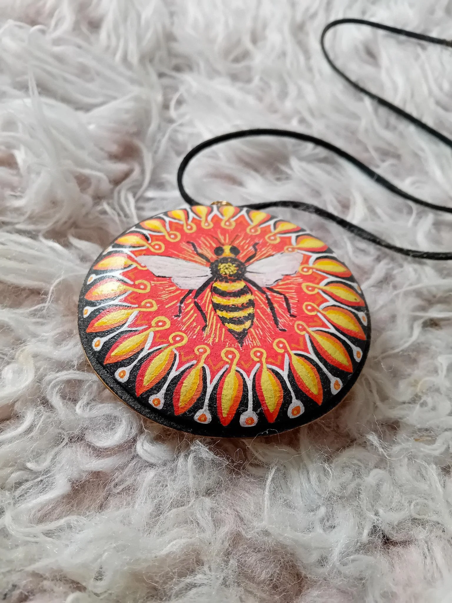 Handgemaakte ronde hanger van balsahout met een geschilderde bij op een gestileerde bloemrijke achtergrond in rood, geel wit en zwart, dat verbonden is met zwart koord en ligt op een witte vacht.