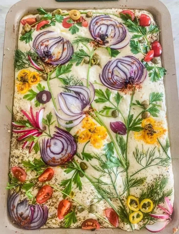 Een grote schaal met een decoratieve salade of een gerecht met gesneden rode uien, kruiden, cherry tomaten, en andere groenten, versierd met bladeren en bloemen.