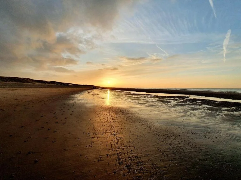 Zonsondergang boven een rustig strand met natte zandbodem en kleine waterplassen, lange schaduwen en een heldere hemel met wolken en strepen.