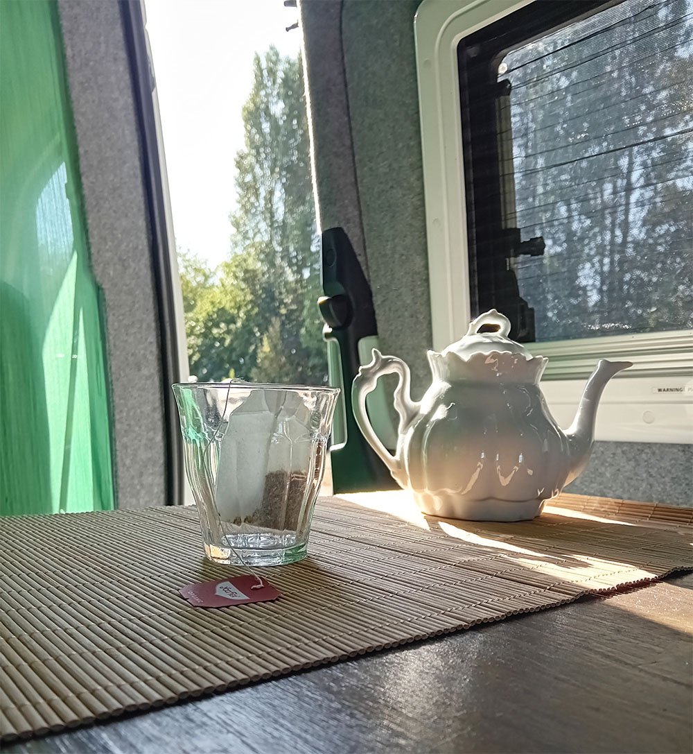 Een houten tafel met een mok koffie en een lepel, in een tuin met gras, struiken en bomen op de achtergrond.