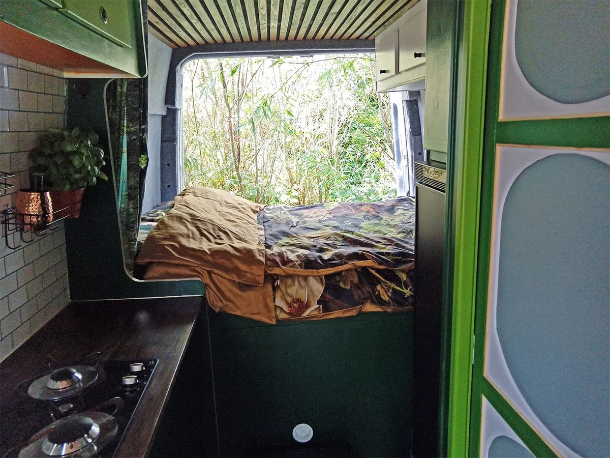 Binnenkant van een camper of kleine kamer met een bed dat bedekt is met een kleurrijk tafelkleed met bloemenpatroon en een wit kussen, een raam met jaloezieën aan de linkerkant en een donker, bekleed plafond.
