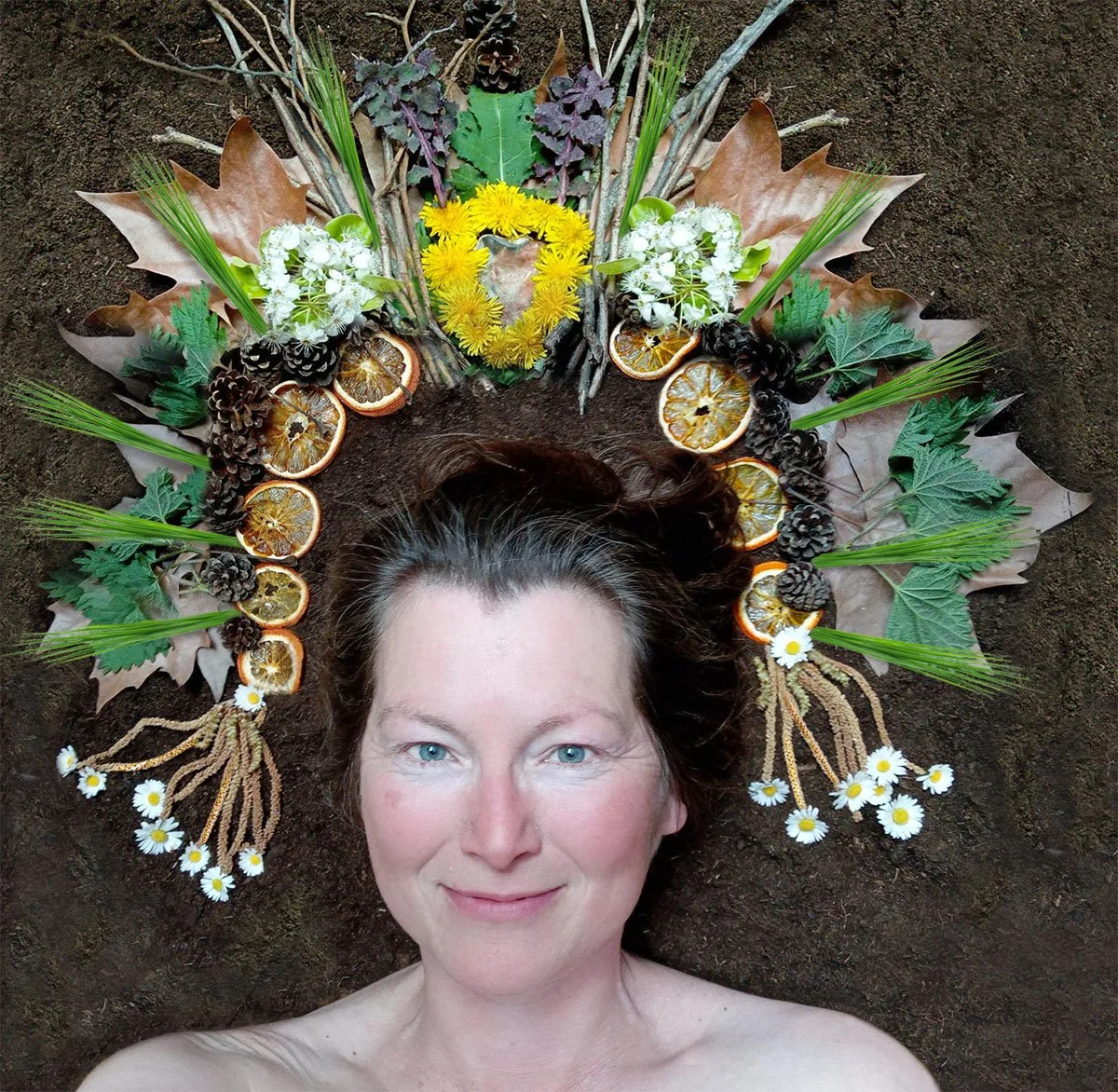 Vrouw ligt met haar hoofd op de grond met een bloemenkrans en natuurlijke decoraties gemaakt van takken, bladeren, gedroogde sinaasappelschijfjes, dennenappels, madeliefjes en gras.