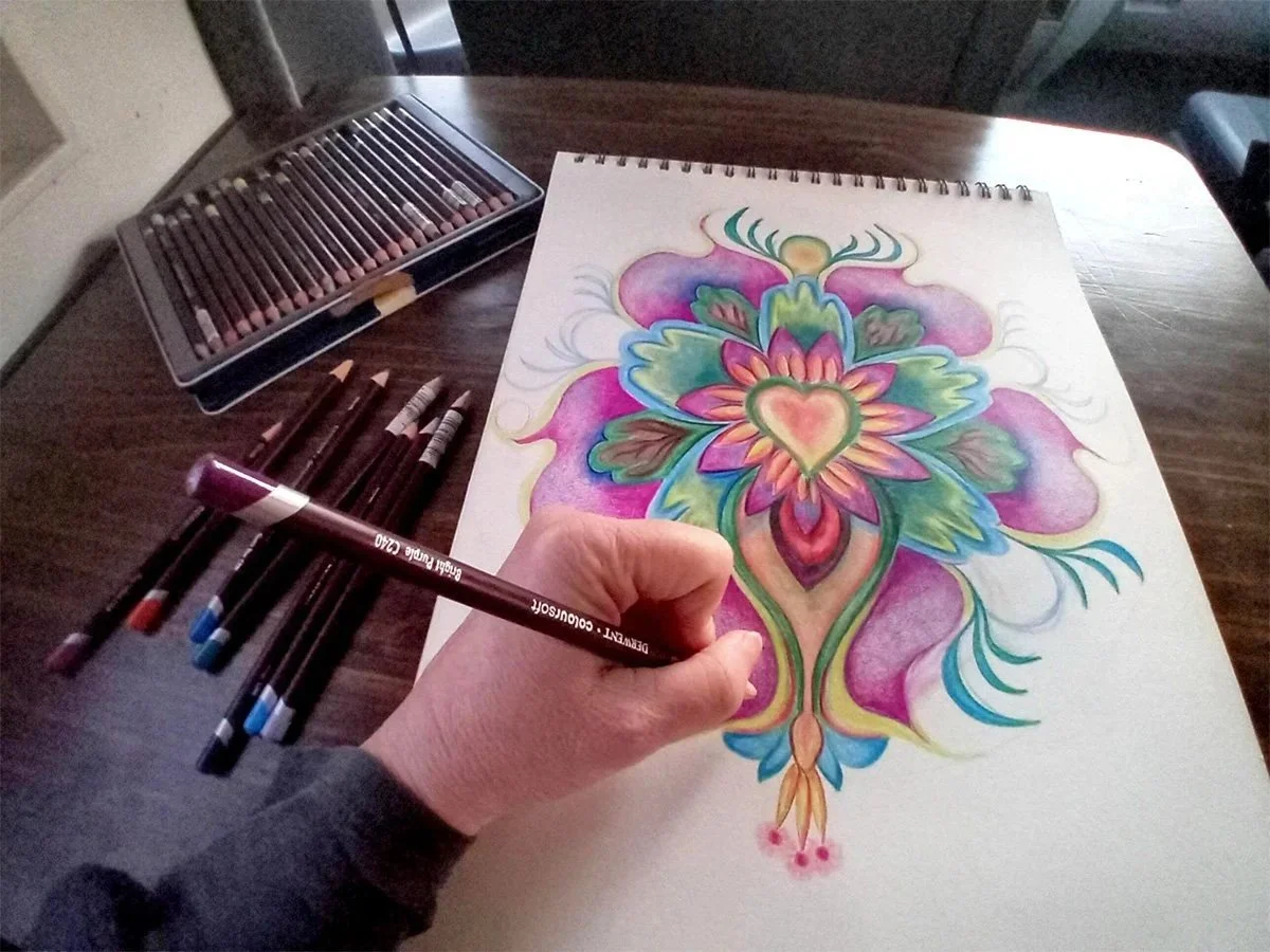 Persoon die een kleurrijk, symmetrisch ontwerp tekent op wit papier, met potloden en stiften op een houten tafel.