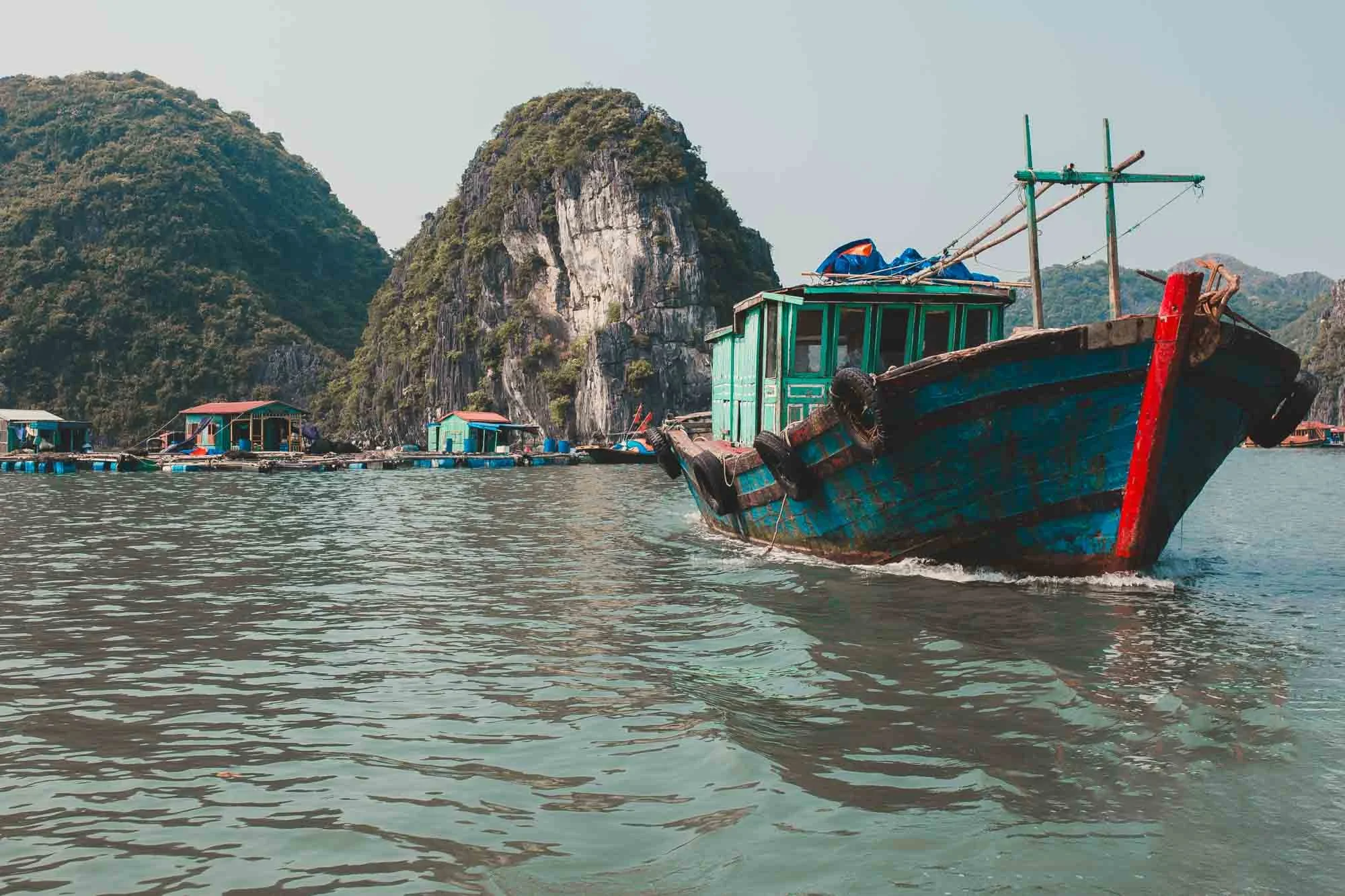 An old wooden boat with green cabin and red accents floating on calm water near limestone cliffs and floating houses.