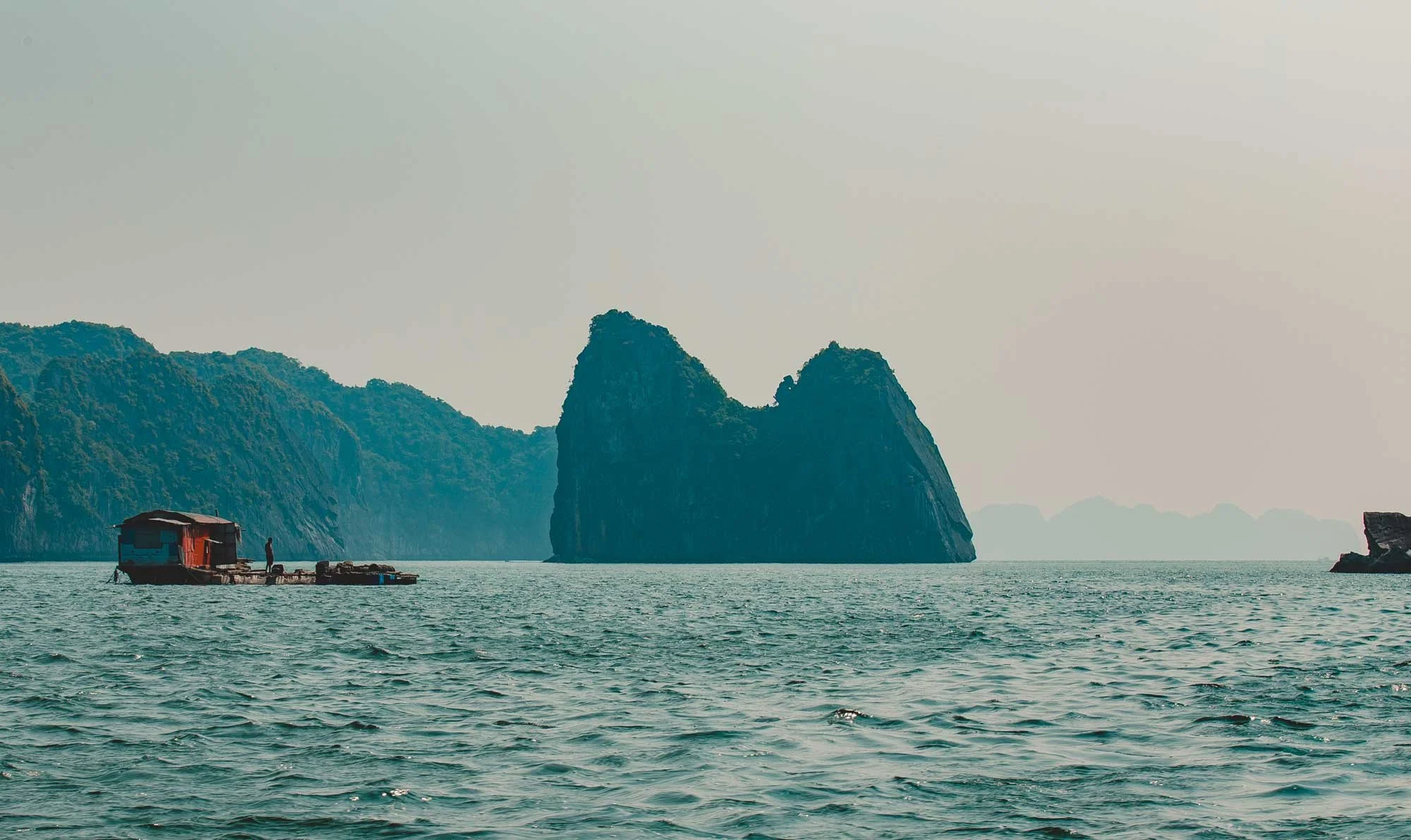 A seascape with a houseboat on the water, a large rocky island formation, and distant landforms under a hazy sky.