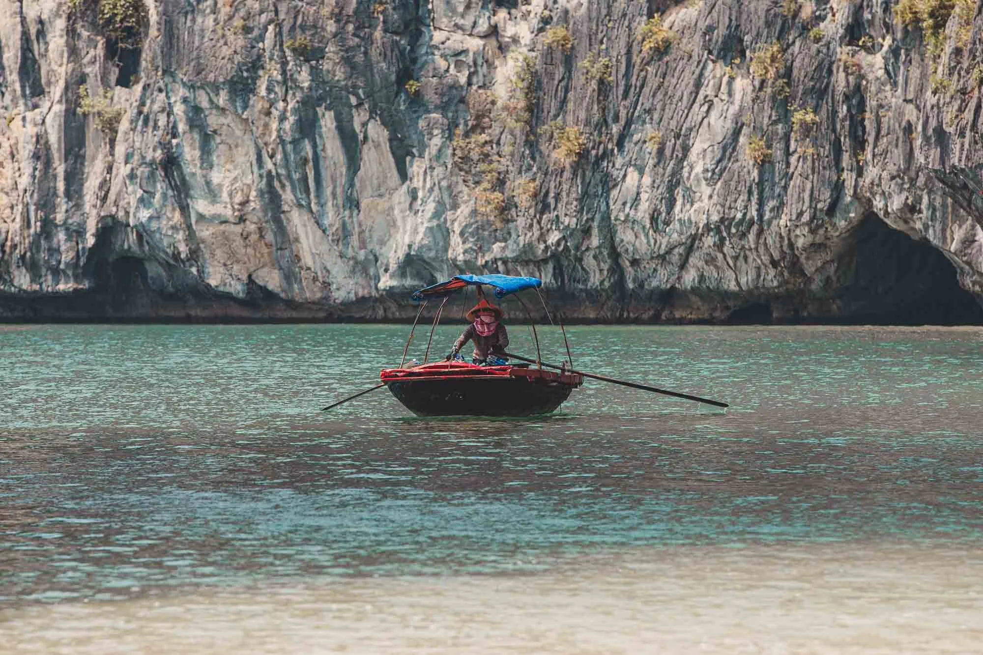 A person in a small boat with a blue canopy rowing on a body of water near a rocky cliff.