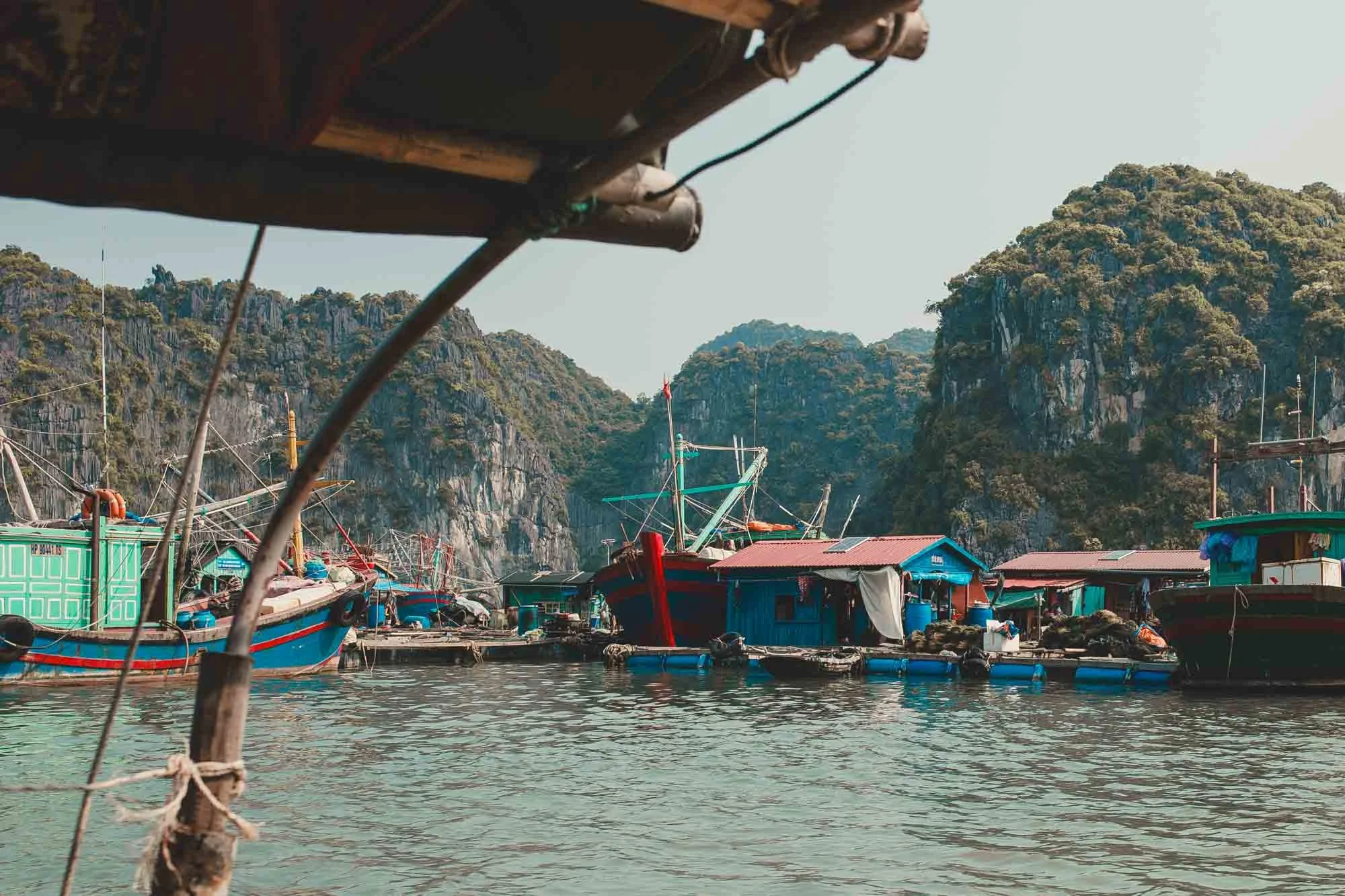 View of boats docked at a floating village with small houses on water, surrounded by lush, green mountains.