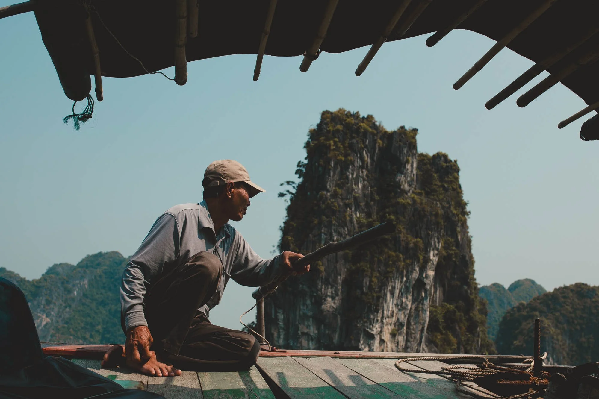 An elderly man sitting on a boat, holding a pole, with limestone karst formations in the background.
