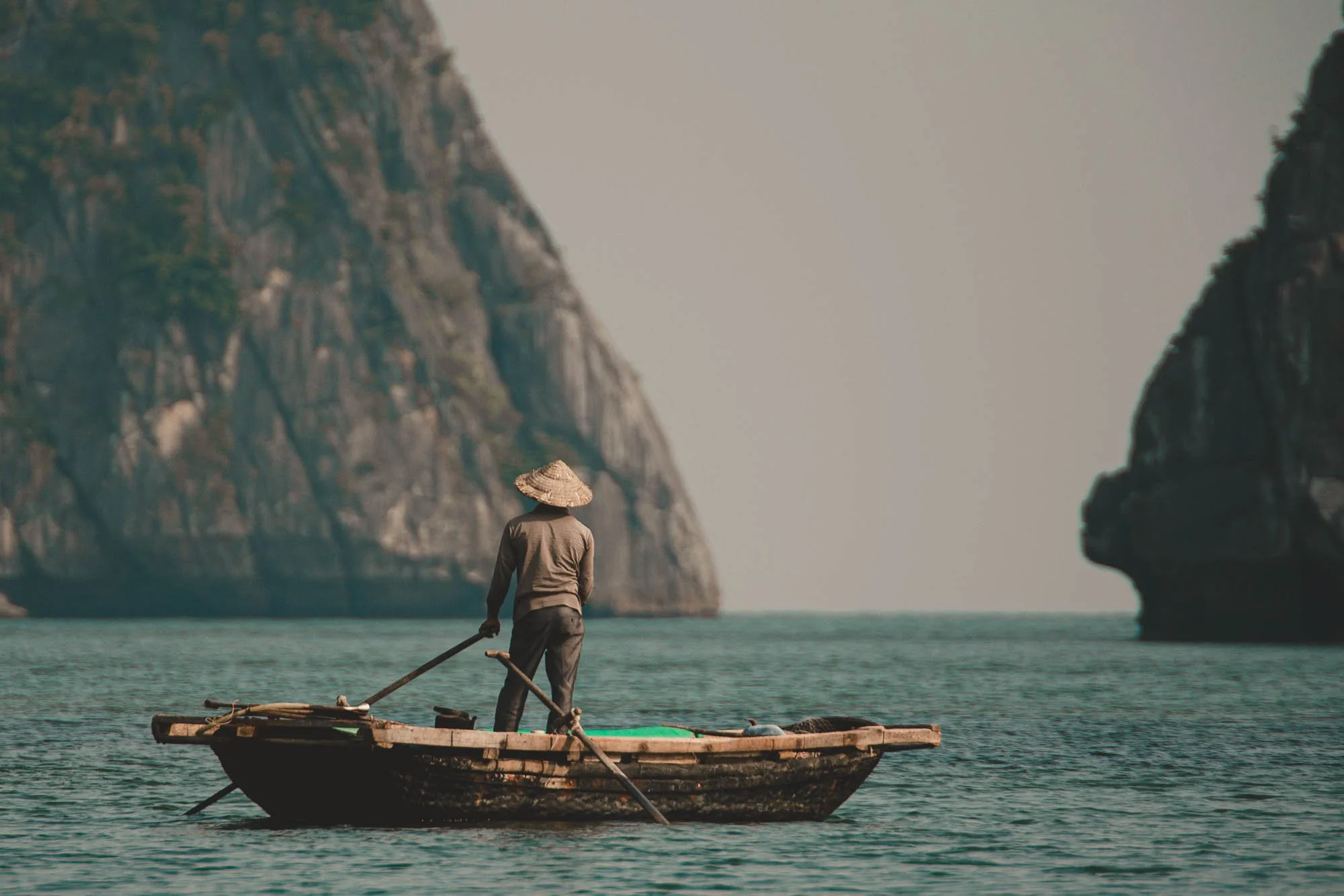 A person wearing a conical hat standing on a small wooden boat in water with large rocky cliffs in the background.