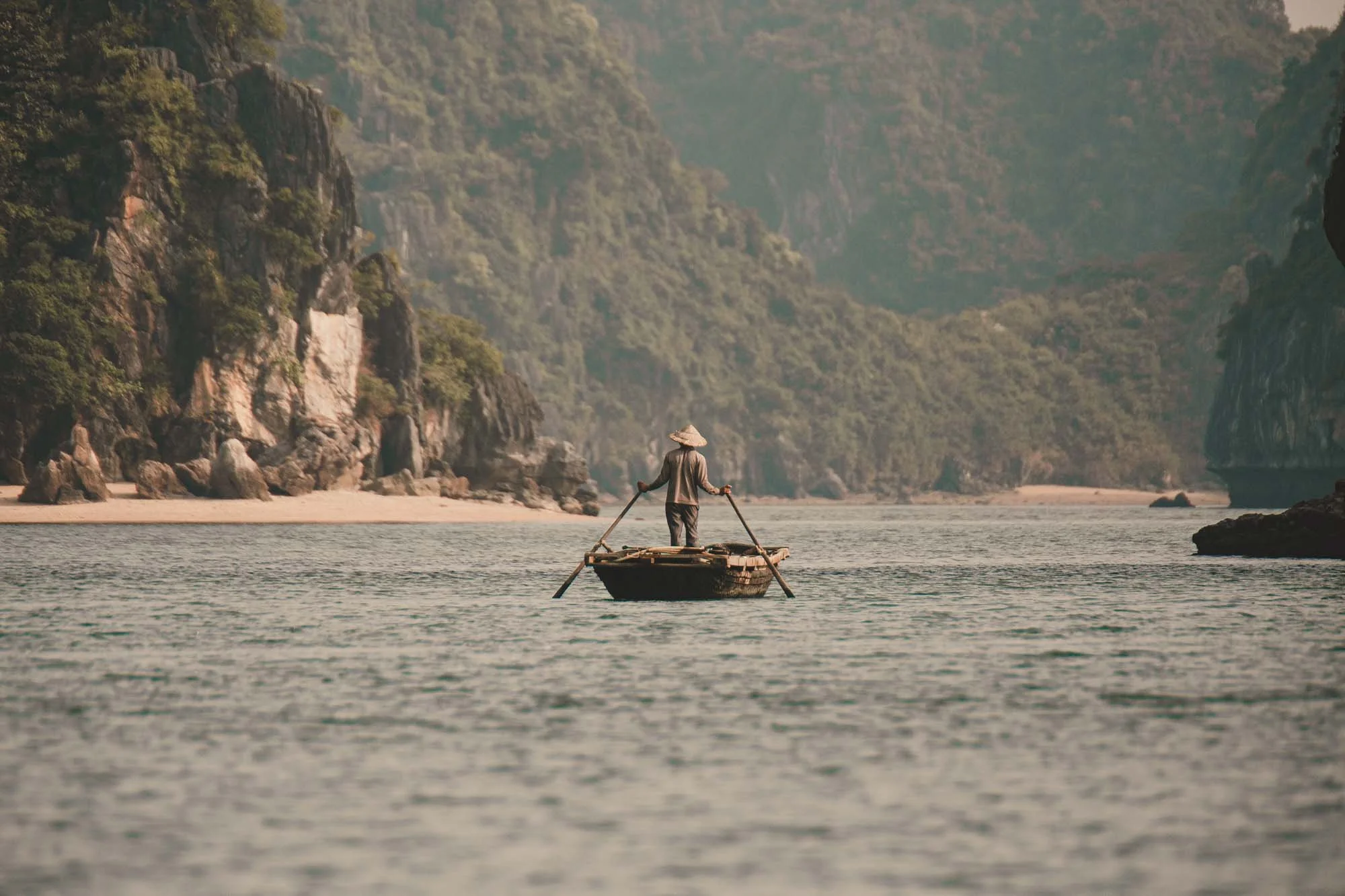 A person wearing a traditional Asian conical hat stands on a small wooden boat in a river surrounded by lush, green cliffs.