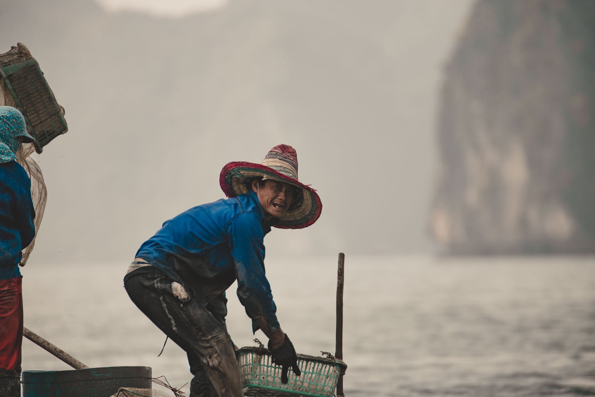 A fisherman wearing a large, colorful straw hat and a blue raincoat, leaning over a fishing net on a boat, with a misty mountain in the background.