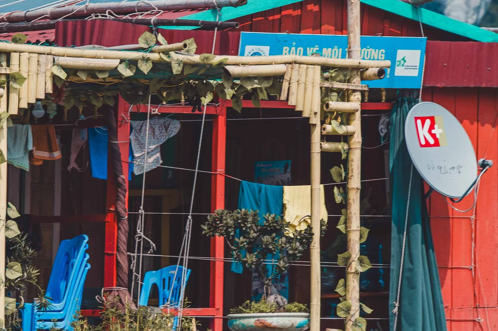 A small outdoor area of a building with bamboo decorations, hanging clothes, and a satellite dish with the K+ logo. There are blue plastic chairs stacked and a potted plant near the entrance.