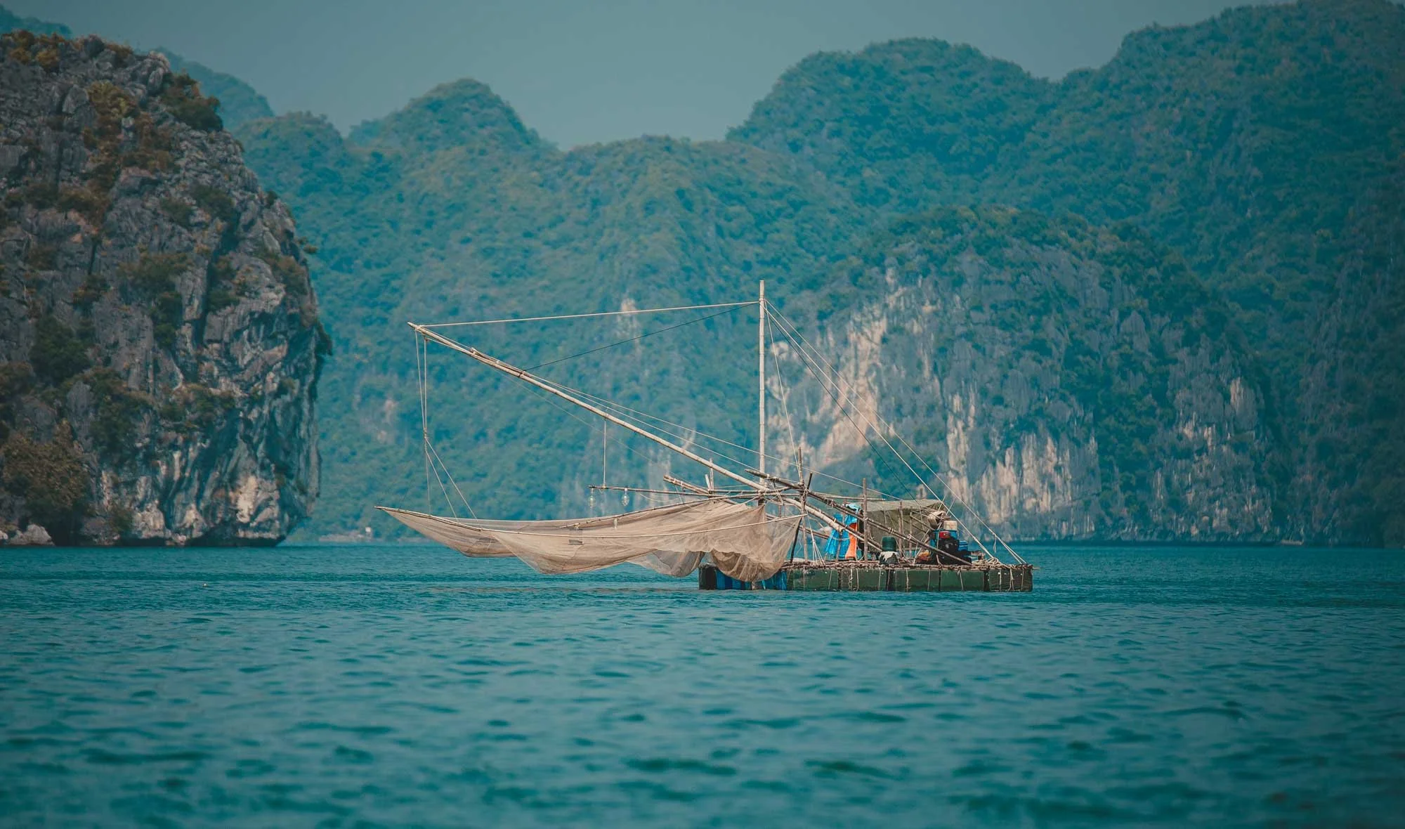 A floating fishing platform with a partially deployed fishing net on a calm blue body of water, surrounded by steep, green, mountainous cliffs.