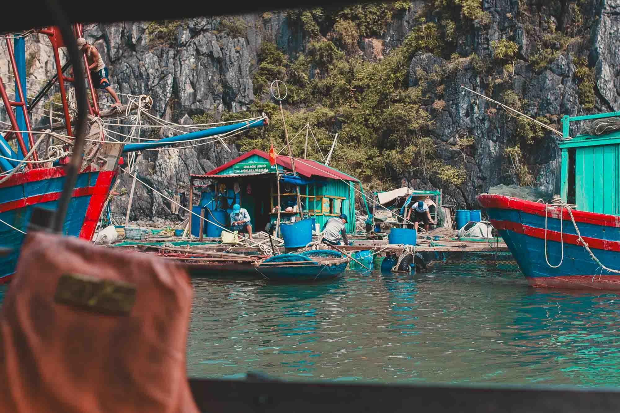 Fishing boats docked at a small floating village with a rocky cliff and greenery in the background.