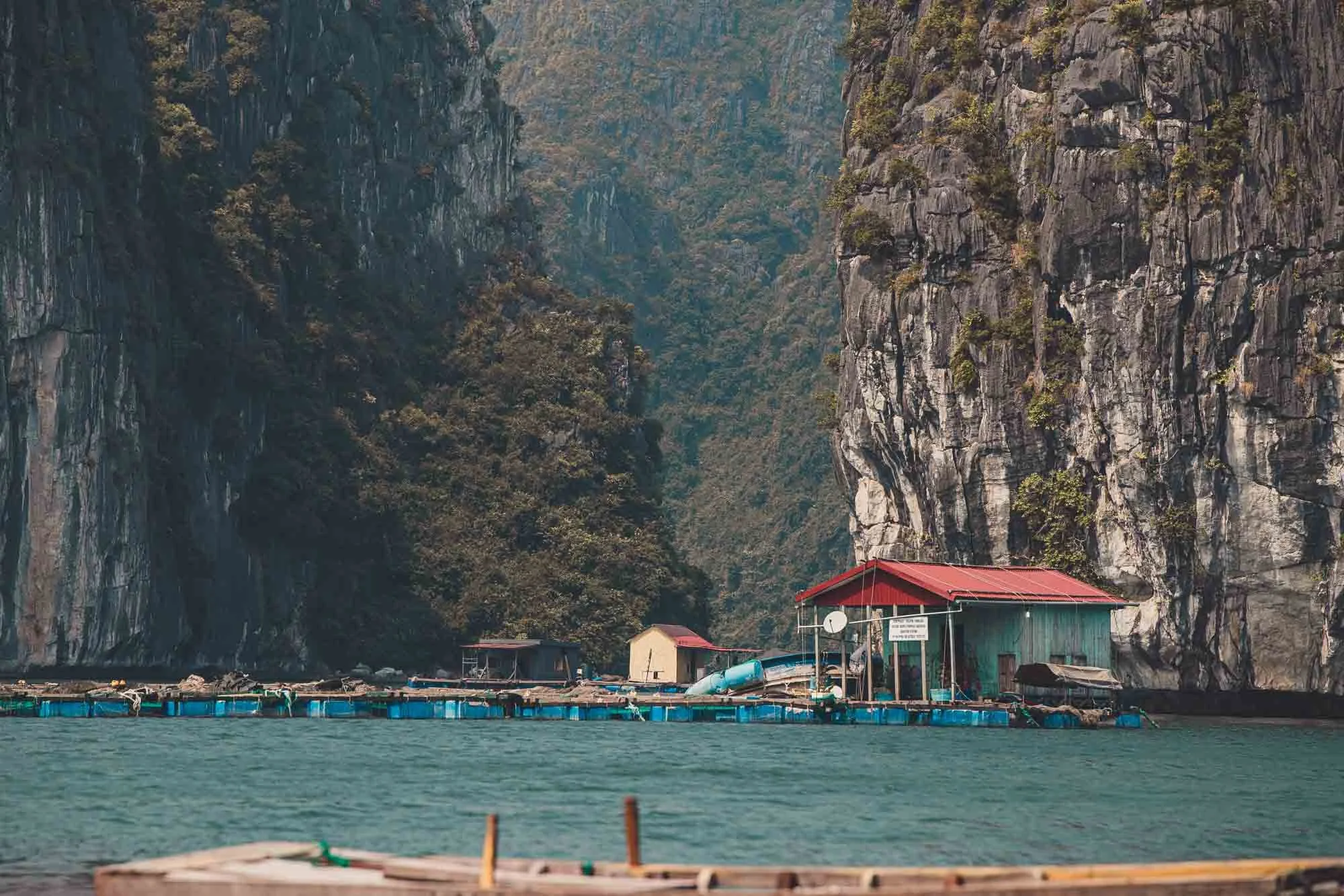 A floating house with a red roof and green walls is situated on the water, surrounded by tall rocky cliffs covered with greenery.