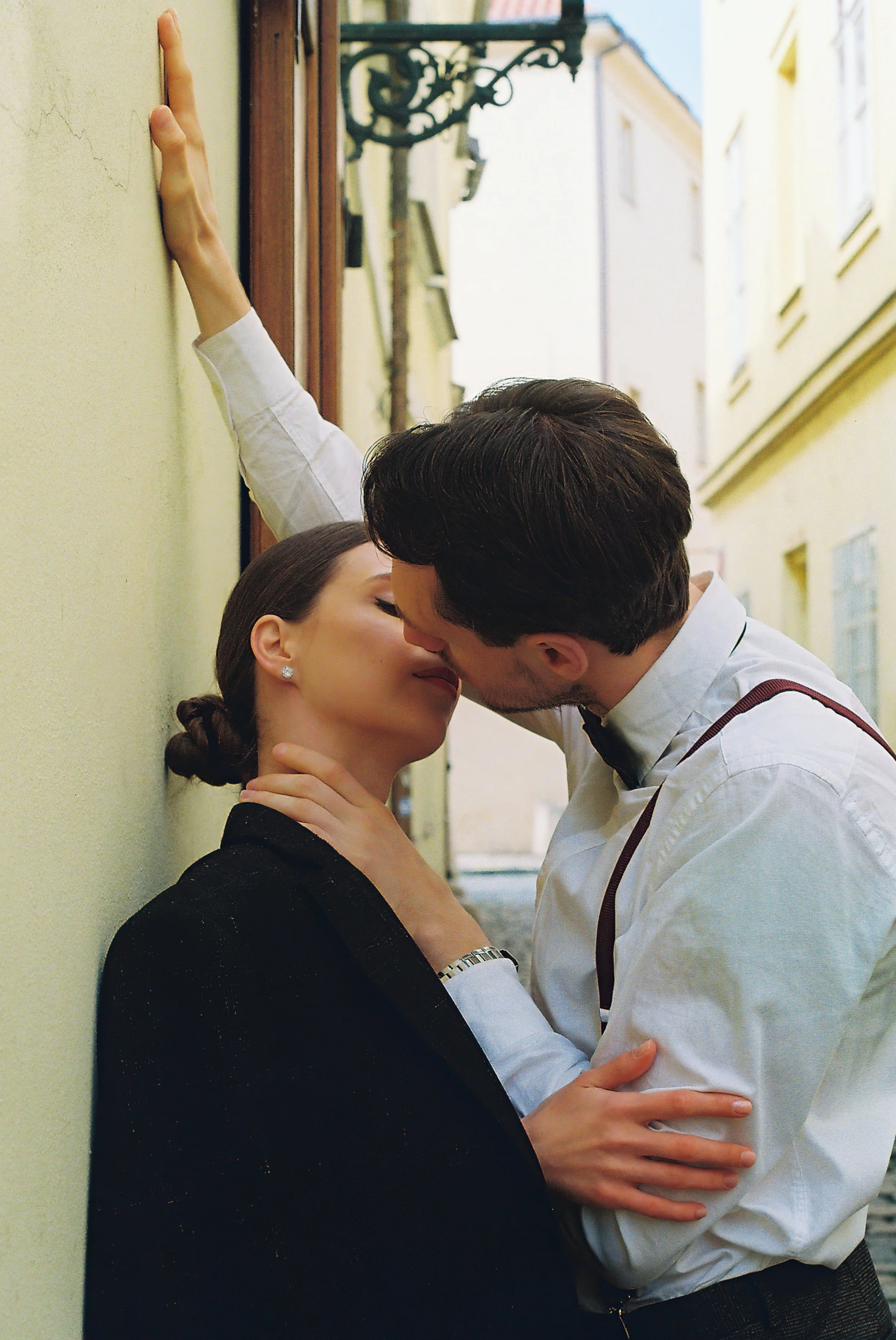Wedding Website Design, Digital Wedding Stationery, Web Design and Branding for Wedding Professionals. A couple kissing against a wall in a narrow street, with the man wearing a white shirt and suspenders, and the woman wearing a dark coat.