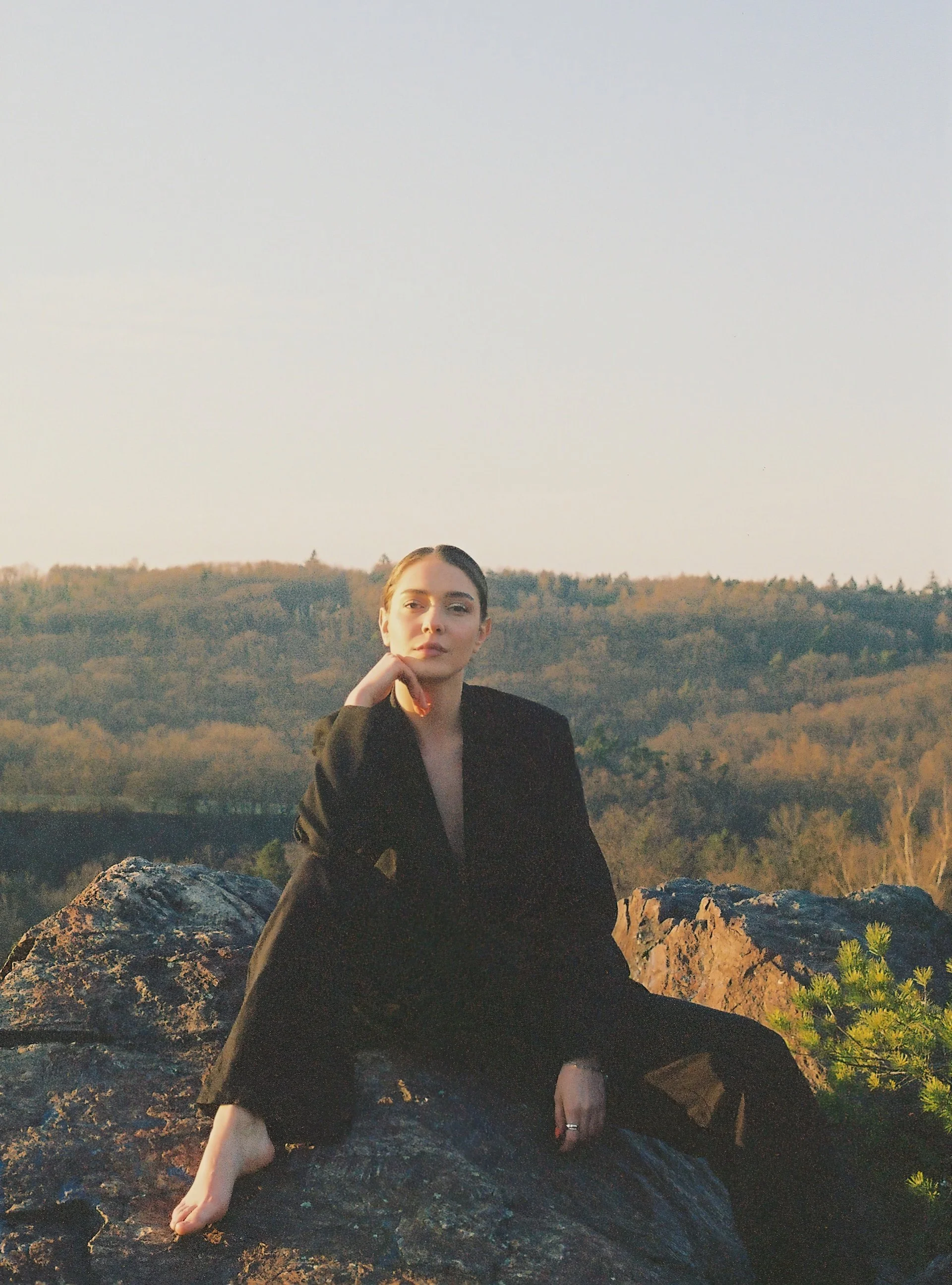 Wedding Website Design, Digital Wedding Stationery, Web Design and Branding for Wedding Professionals | Avelã White. Person in black suit sitting on a rock with a scenic view of a forest and hills in the background, under a clear sky.