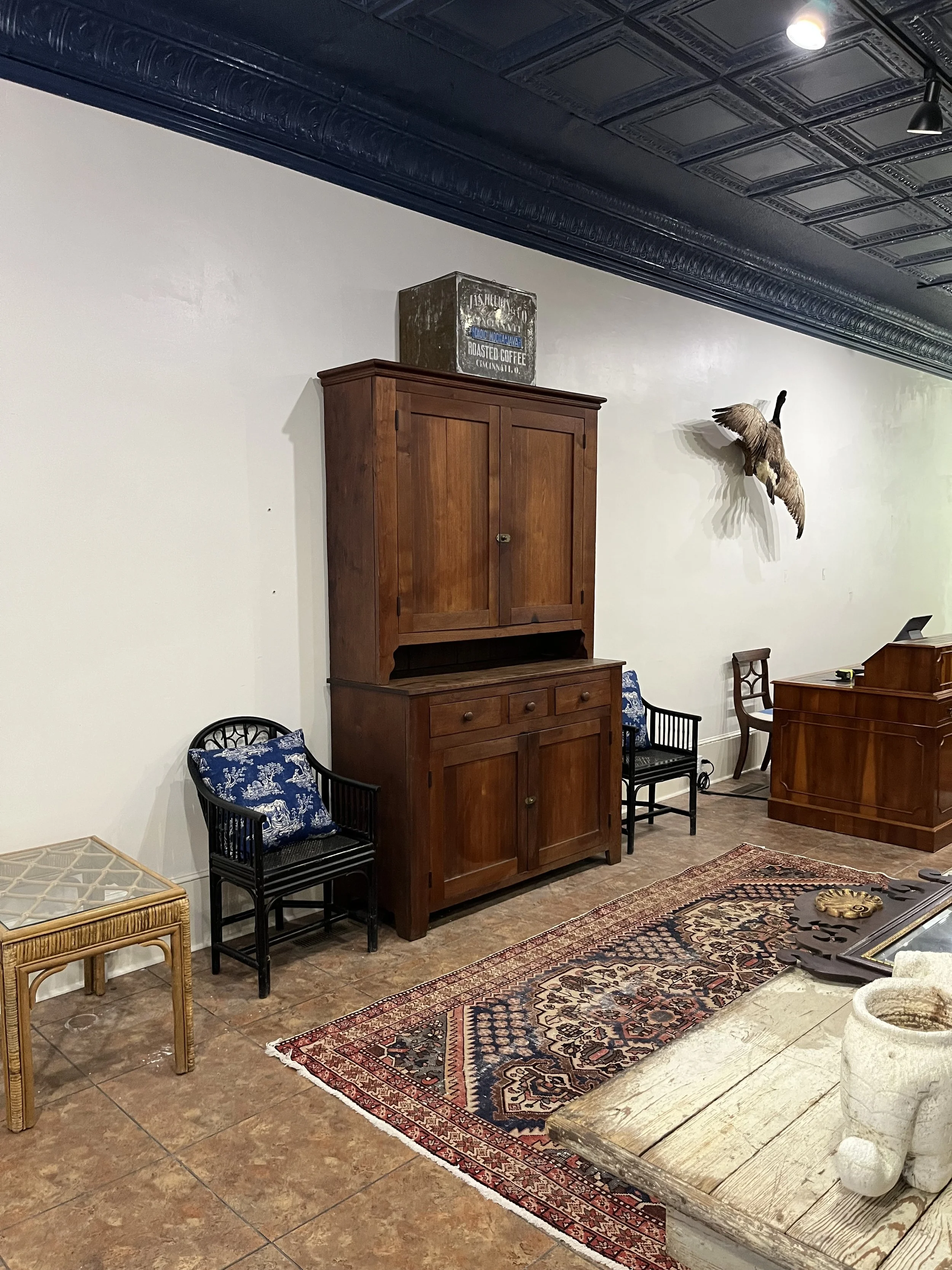 A vintage-style room with a wooden cabinet, two black chairs with blue patterned cushions, a colorful patterned rug, a wooden desk or counter, and a mounted bird on the wall.