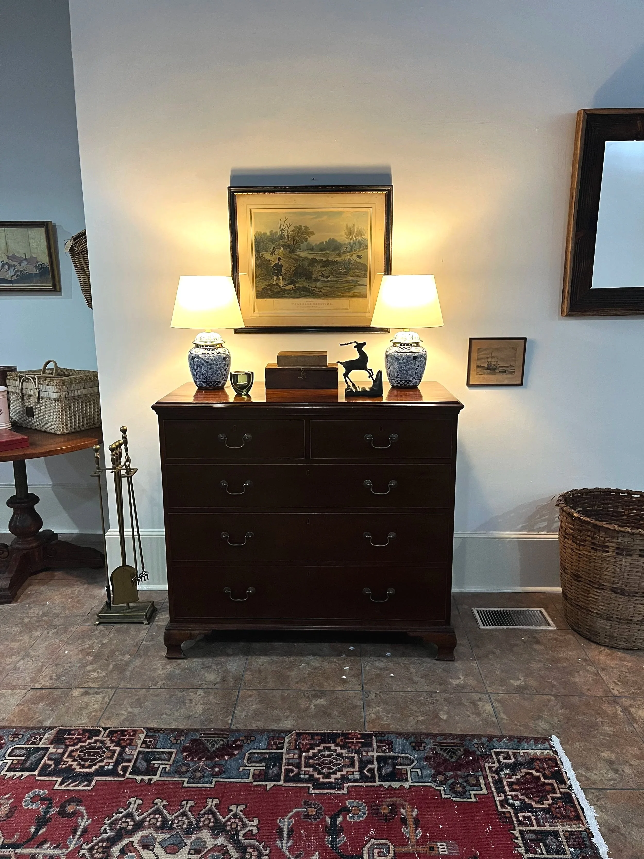 A dark wooden dresser with six drawers, topped with two table lamps, a framed picture above, and decorative items including a sculpture of a rabbit, a small box, and a candle holder. A framed picture is on the wall next to it, and a woven basket is on the floor.