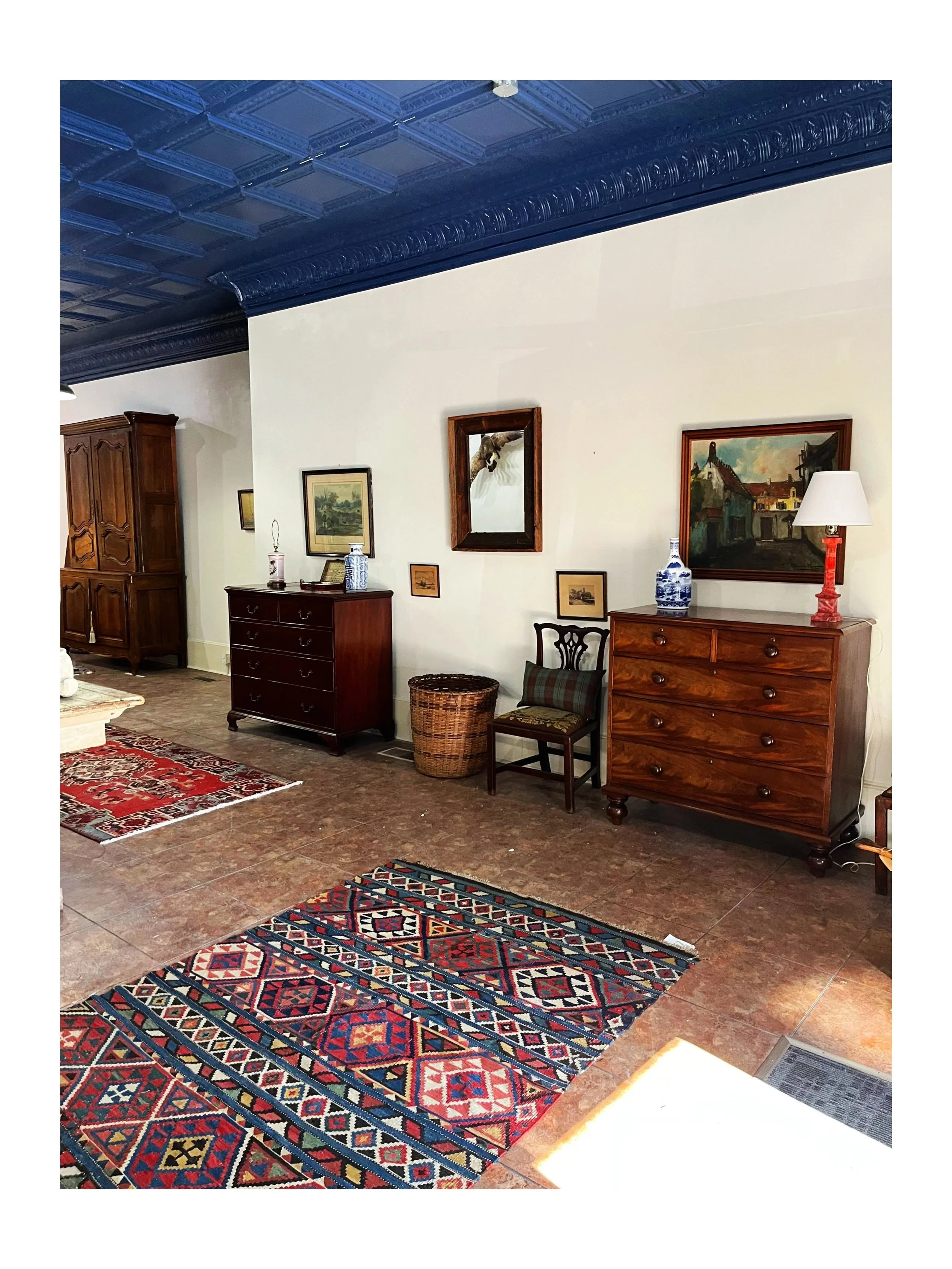 A room with vintage furniture, including a dark wood dresser and a tall cabinet, decorated with framed artwork and vases. Rug with intricate geometric patterns and a planted basket on a tiled floor. The ceiling is painted dark blue with ornate detailing.