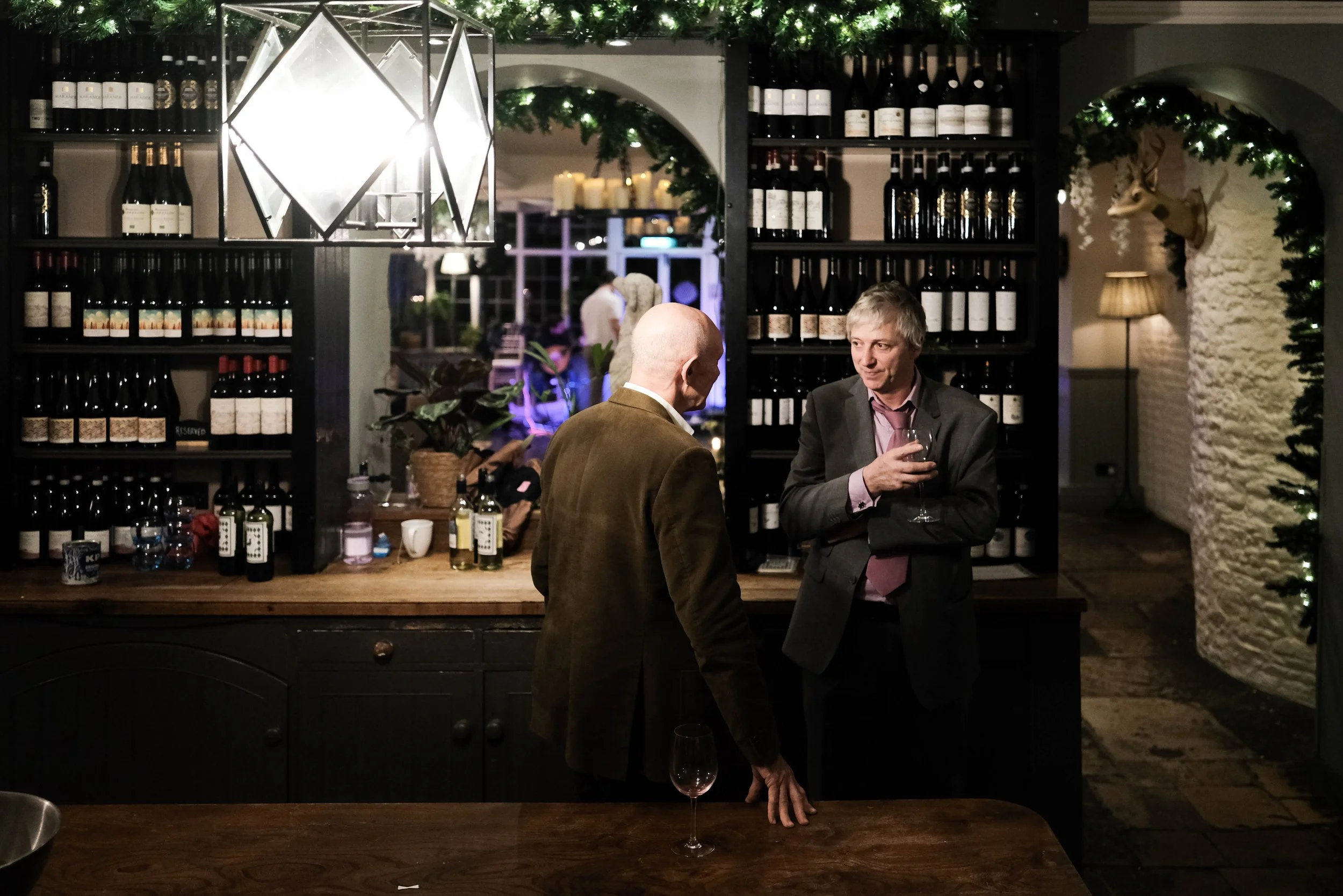 Two men talking at a wine bar, one with glasses of wine, shelves of wine bottles behind them, decorated with greenery, in a warmly lit cozy setting.