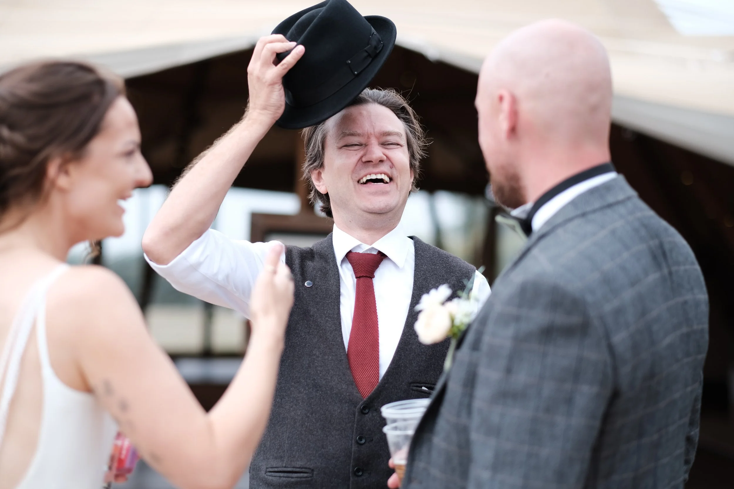 Three people at a wedding celebration, one man smiling and holding a black hat, woman with long hair smiling, man in a suit with a boutonniere.