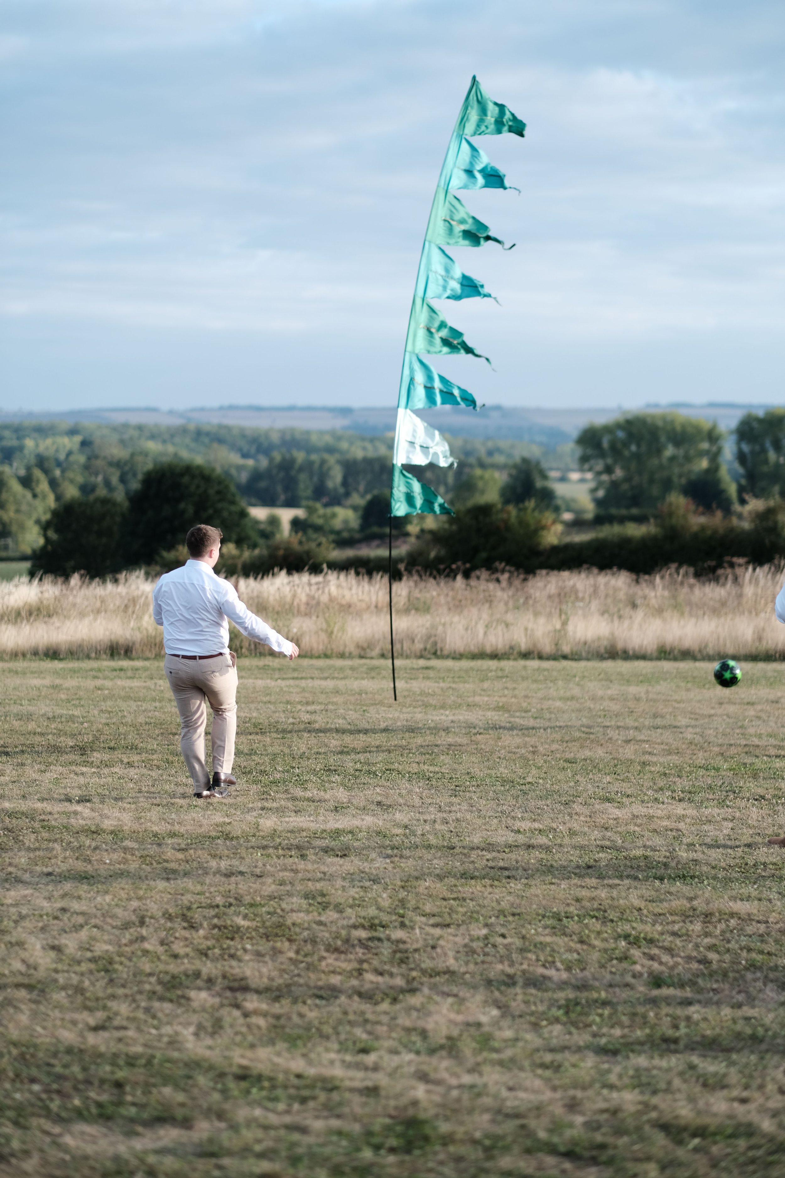 A man in a white shirt and tan pants walking on a grassy field near a green flag on a pole, with a soccer ball nearby and a scenic landscape with trees and hills in the background on a cloudy day.