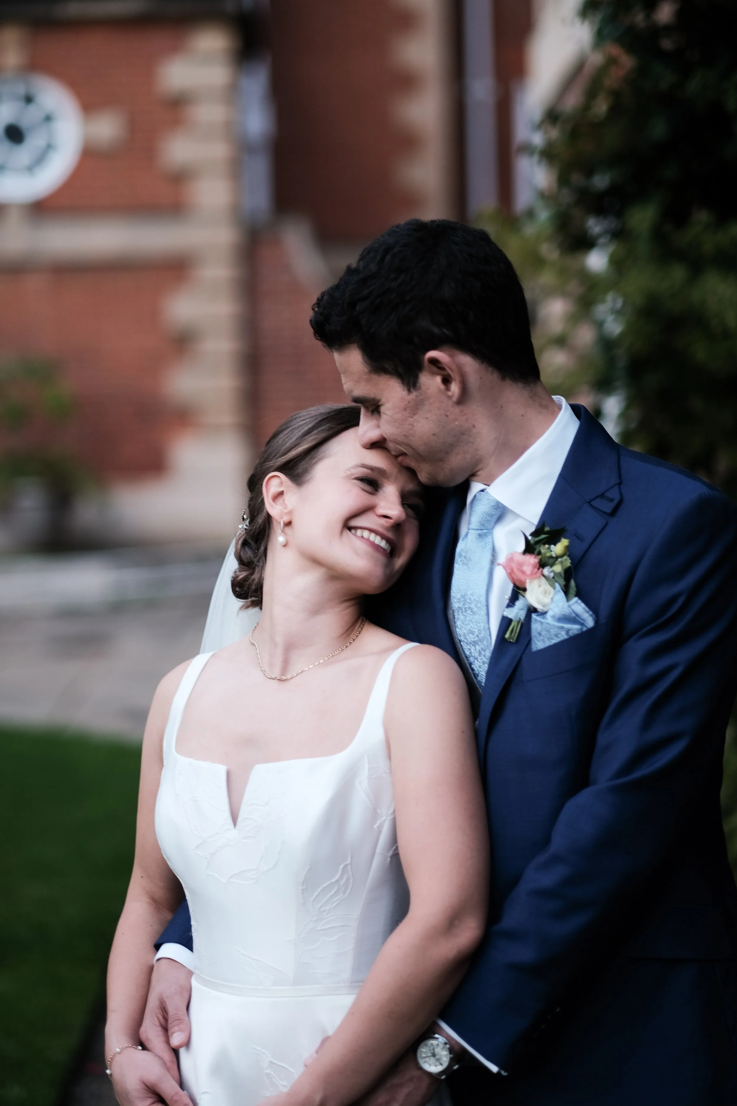 A bride and groom sharing a loving moment outdoors, with the groom gently kissing the bride's forehead. The bride is smiling happily, dressed in a sleeveless white wedding gown, and the groom is wearing a navy suit with a light blue tie and boutonnie
