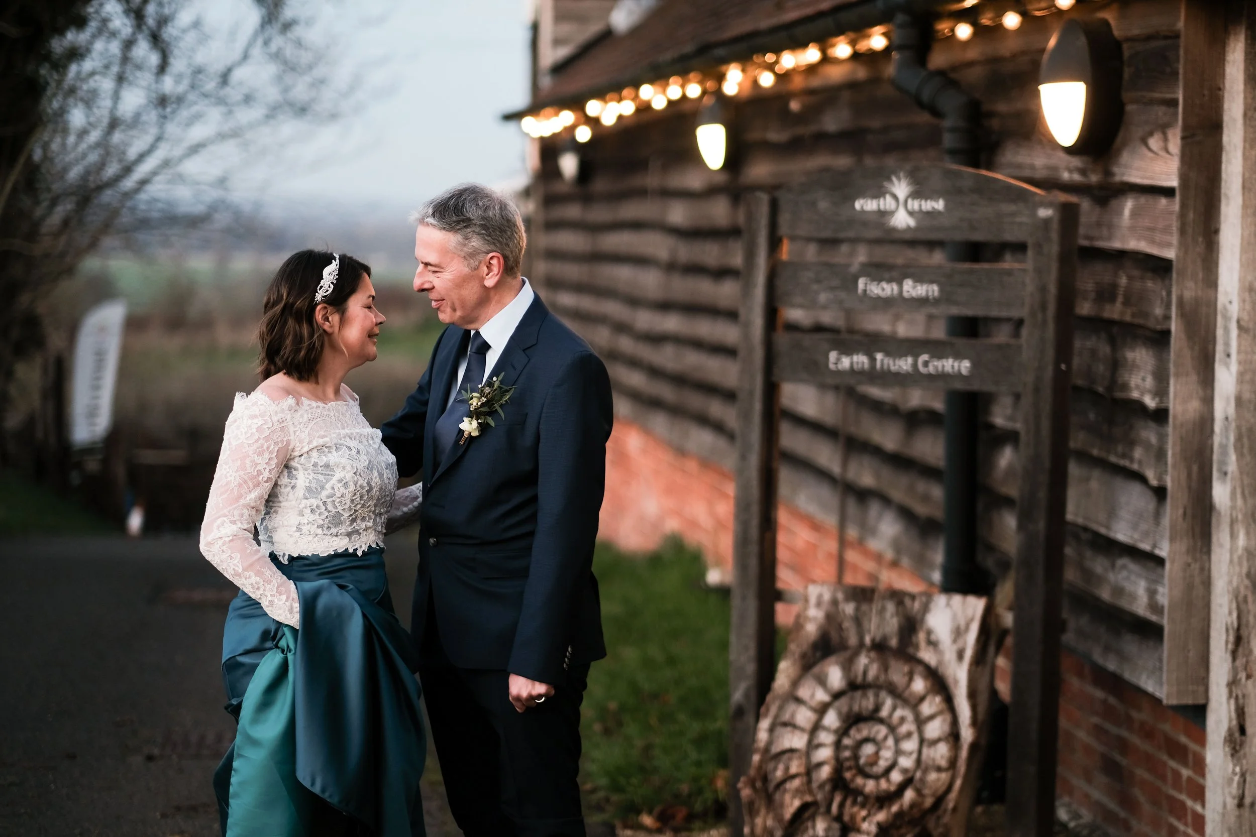 A bride and groom sharing a joyful moment outdoors, standing close and smiling at each other in front of a rustic wooden building with string lights, a sign indicating "Earth Trust Centre" and "Fison Barn."